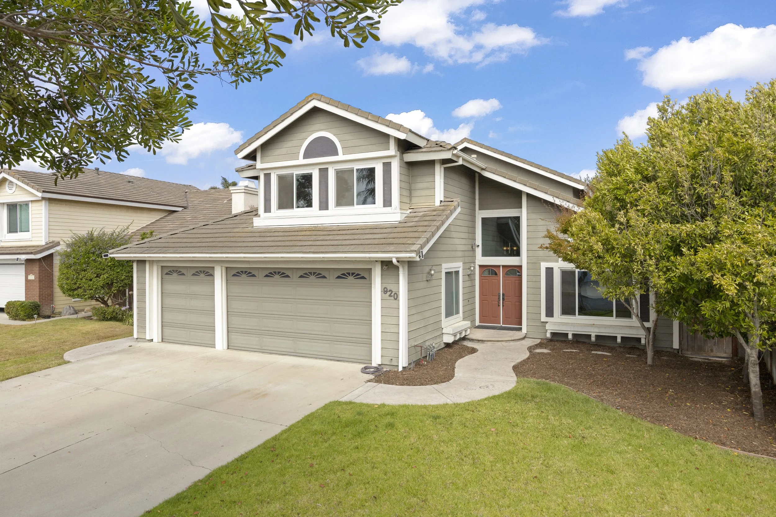 A two-story suburban house with gray siding, a front porch with a double wooden door, and a three-car garage. The front yard has a concrete driveway, a small curved walkway, a green lawn, and trees on the right side. The house has a gabled roof, and 