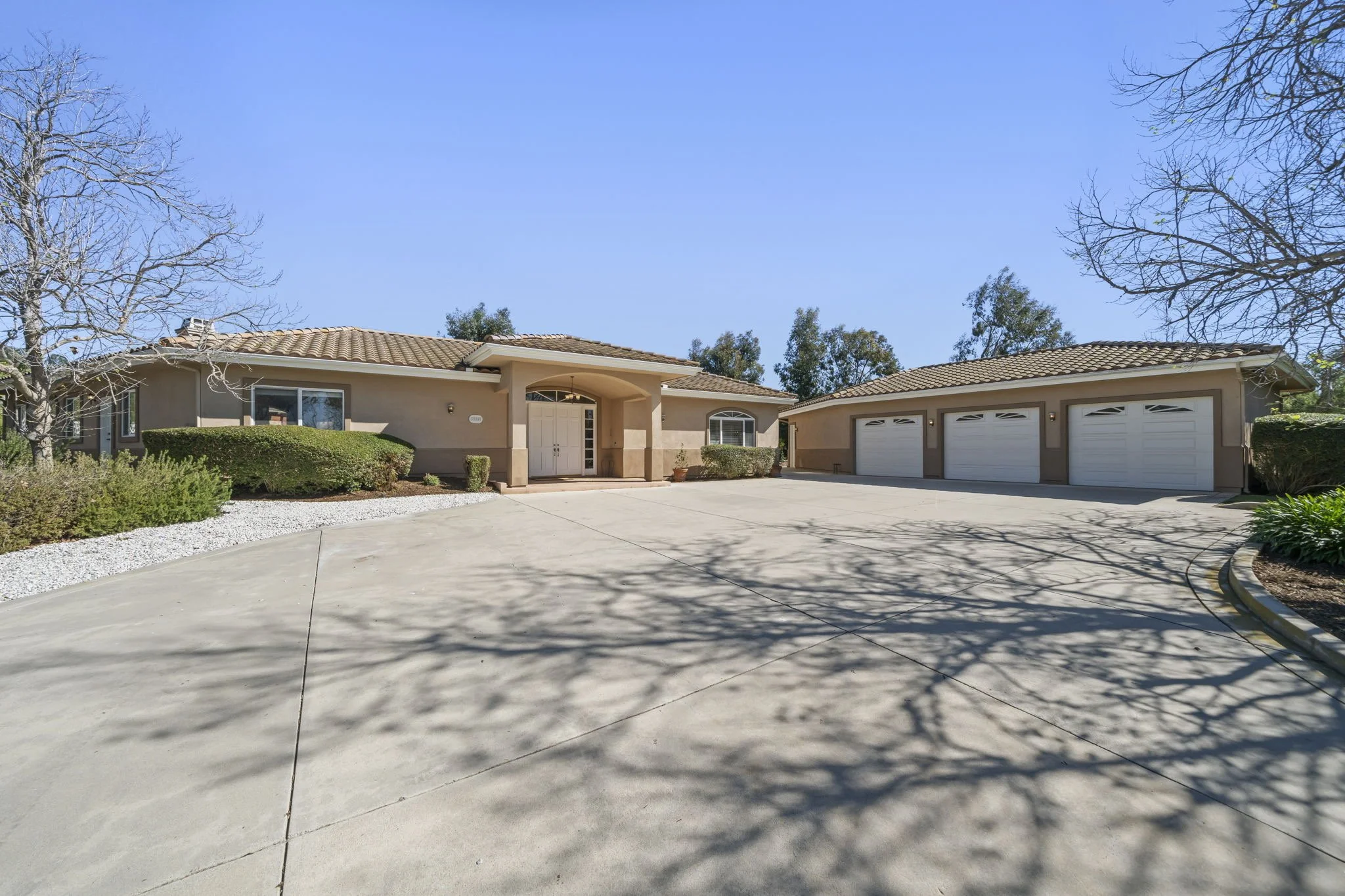 Single-story house with three-car garage and a curved driveway, surrounded by trees and bushes, under a clear blue sky.