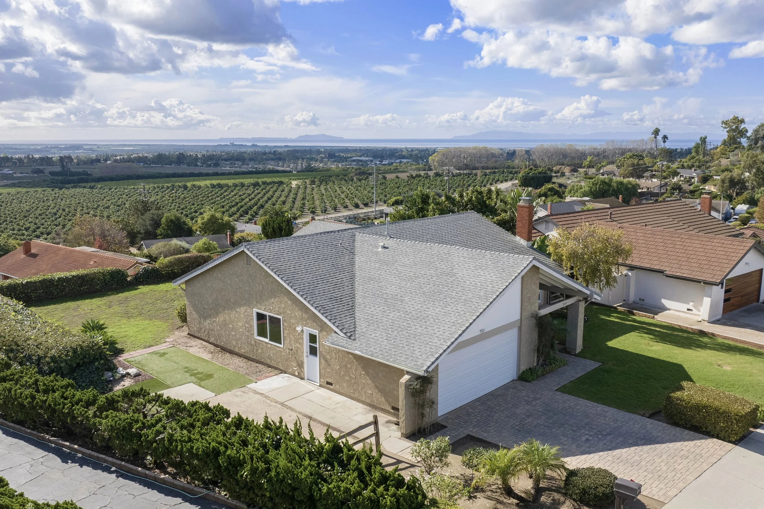 A house with a beige exterior, gray roof, and white garage, surrounded by greenery and neighboring houses, with a view of farmland and blue sky in the background.