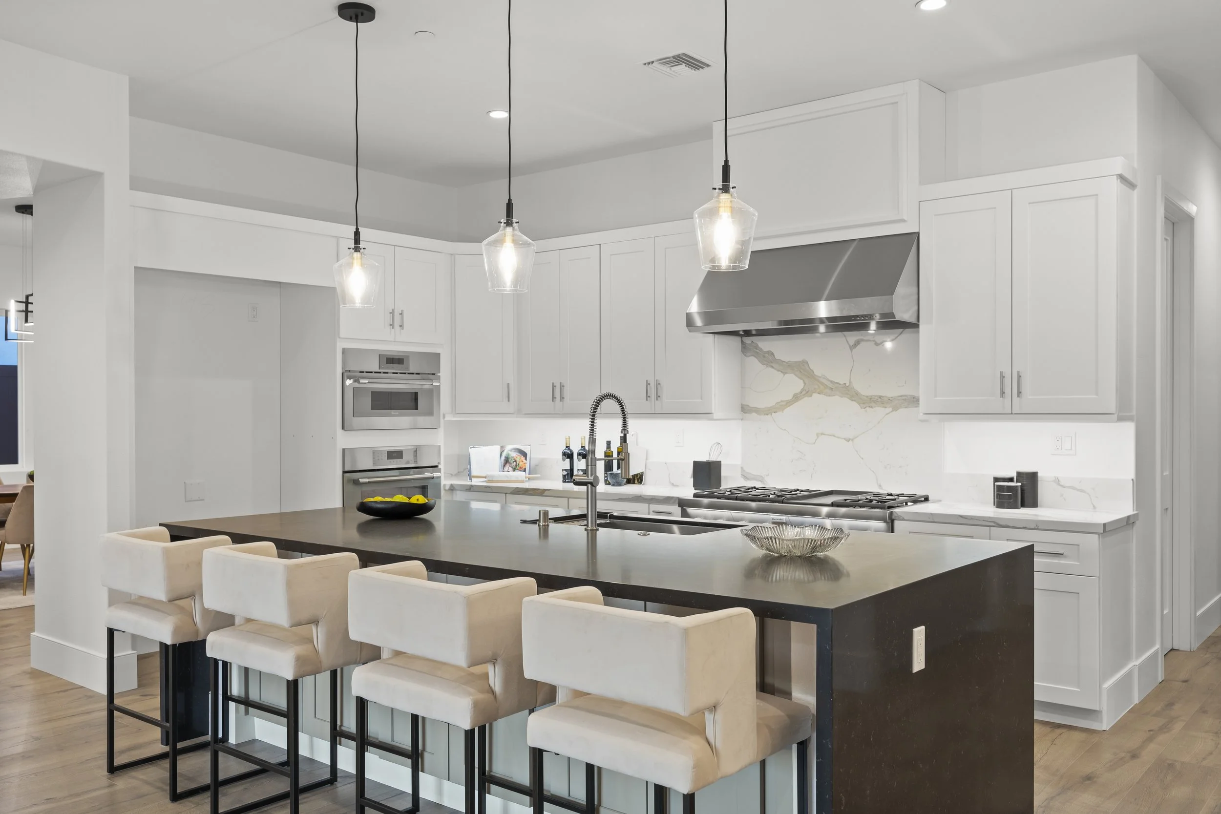 Modern kitchen with white cabinets, marble backsplash, stainless steel appliances, and a large black island with beige stools and pendant lights hanging above.