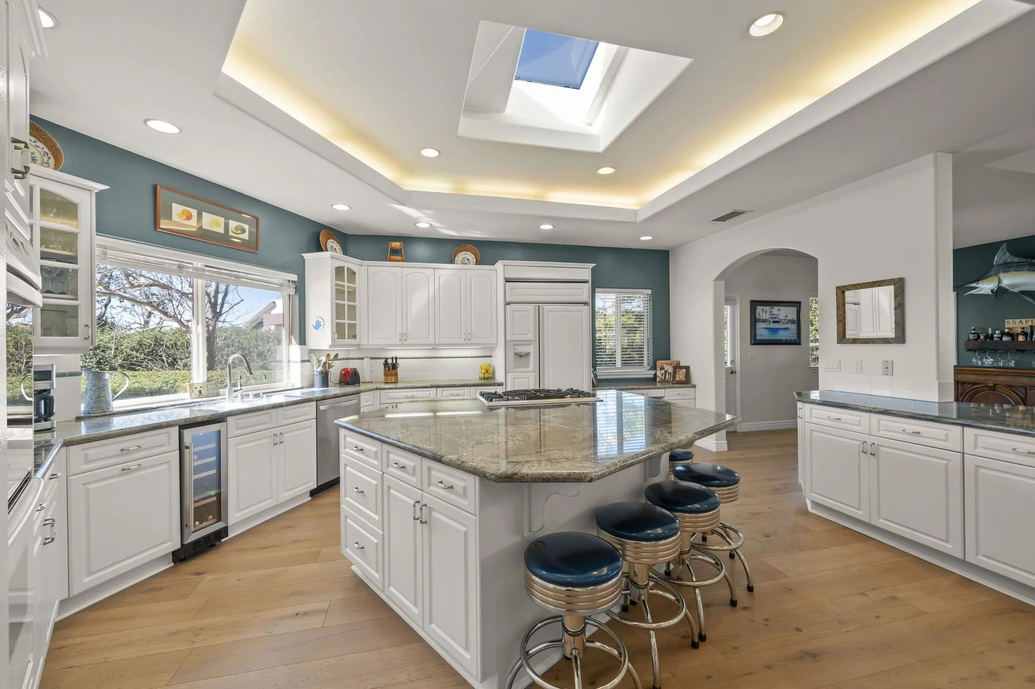 Modern kitchen with white cabinetry, a large granite island with four bar stools, hardwood floors, skylight, and windows showing outdoor greenery.