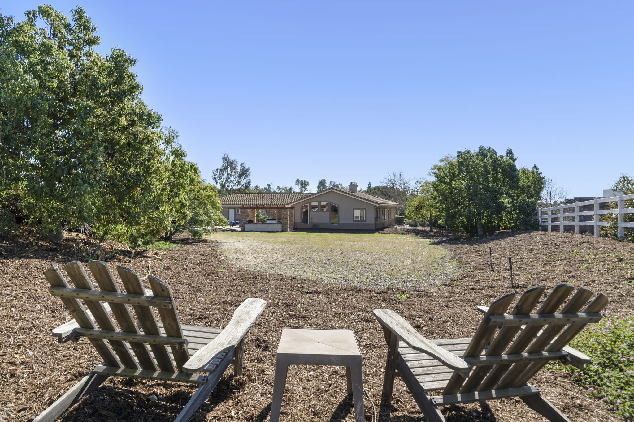 View of a backyard with two wooden chairs and a small table, surrounded by trees and a house in the distance on a clear, sunny day.