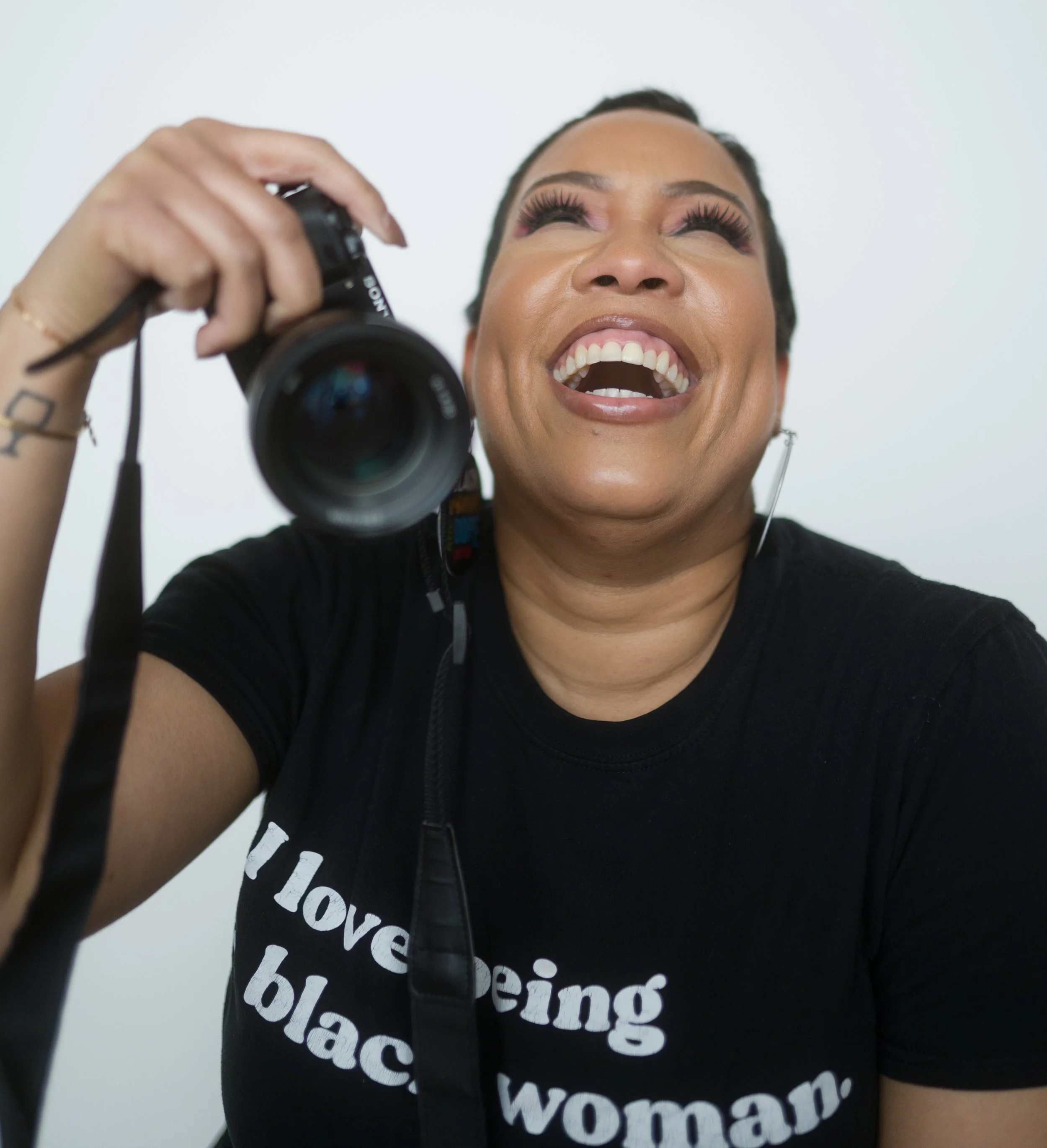 A woman laughing while holding a camera, wearing a black shirt that says I love being a black woman, against a plain white background.