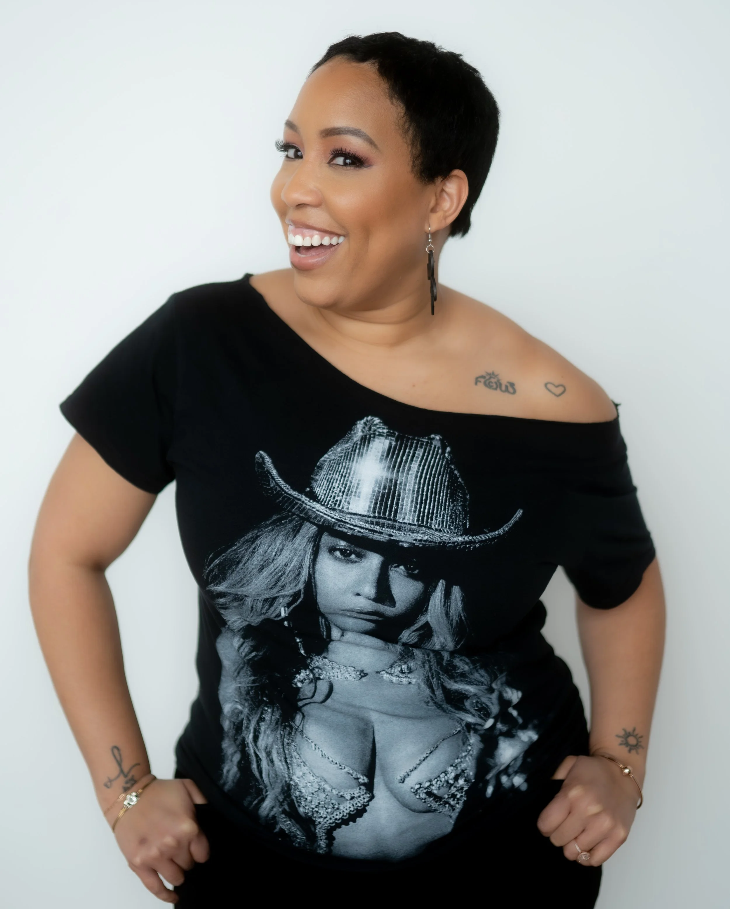 Woman with short black hair smiling, wearing a black off-the-shoulder T-shirt featuring a woman with a cowboy hat. She has tattoos on her shoulder and wrist, is accessorized with earrings, and is posing with her hands on her hips against a plain white background.
