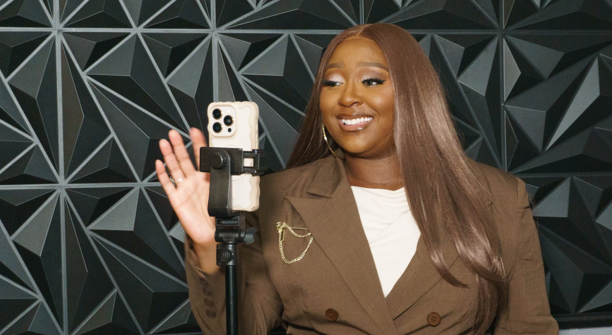 A woman with long brown hair wearing a brown blazer and white shirt, smiling and waving at a smartphone mounted on a stand, against a black geometric patterned wall.