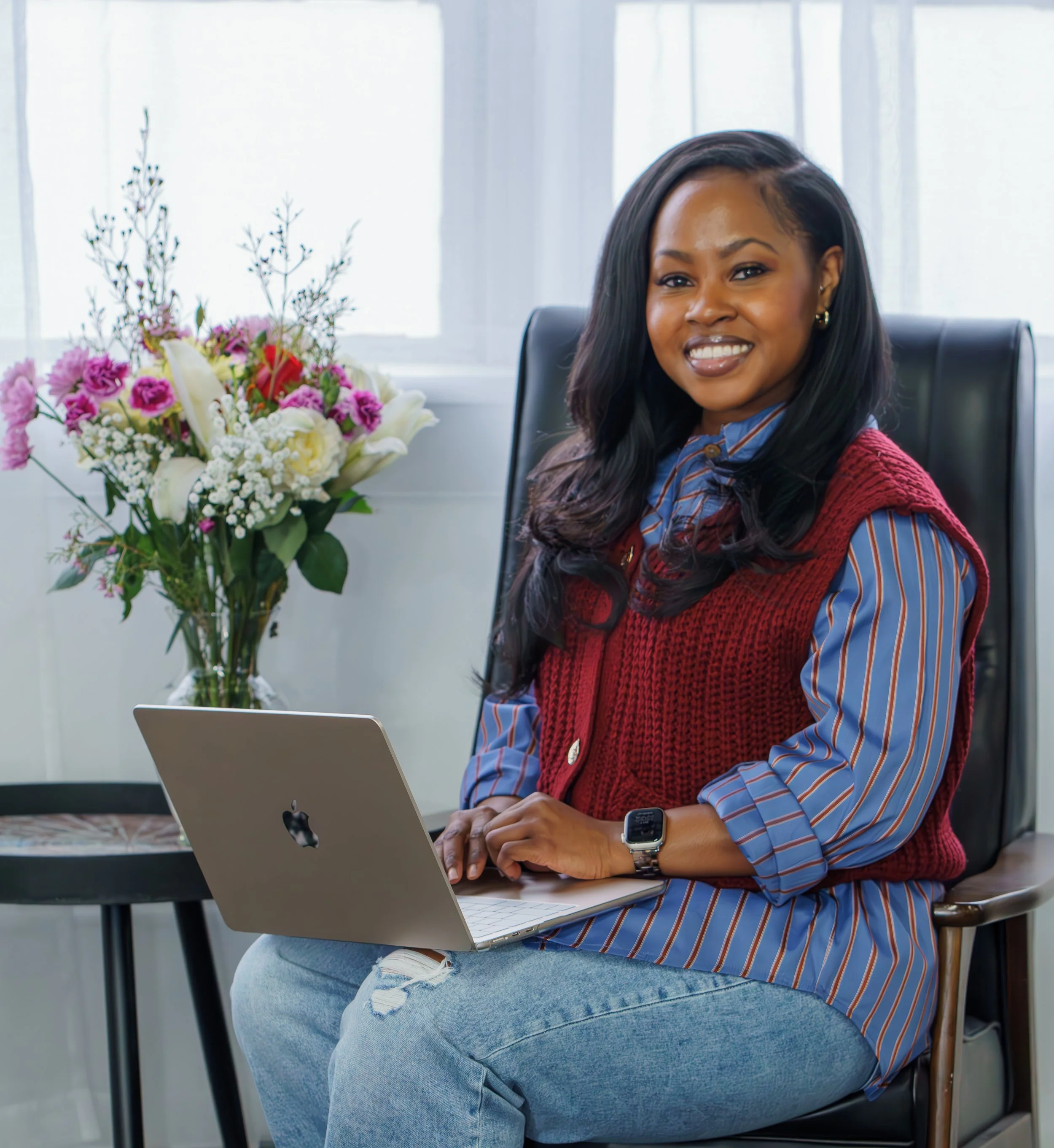 A woman sitting at a desk with a MacBook laptop, smiling, with a bouquet of flowers on a side table beside her, in a bright office setting.