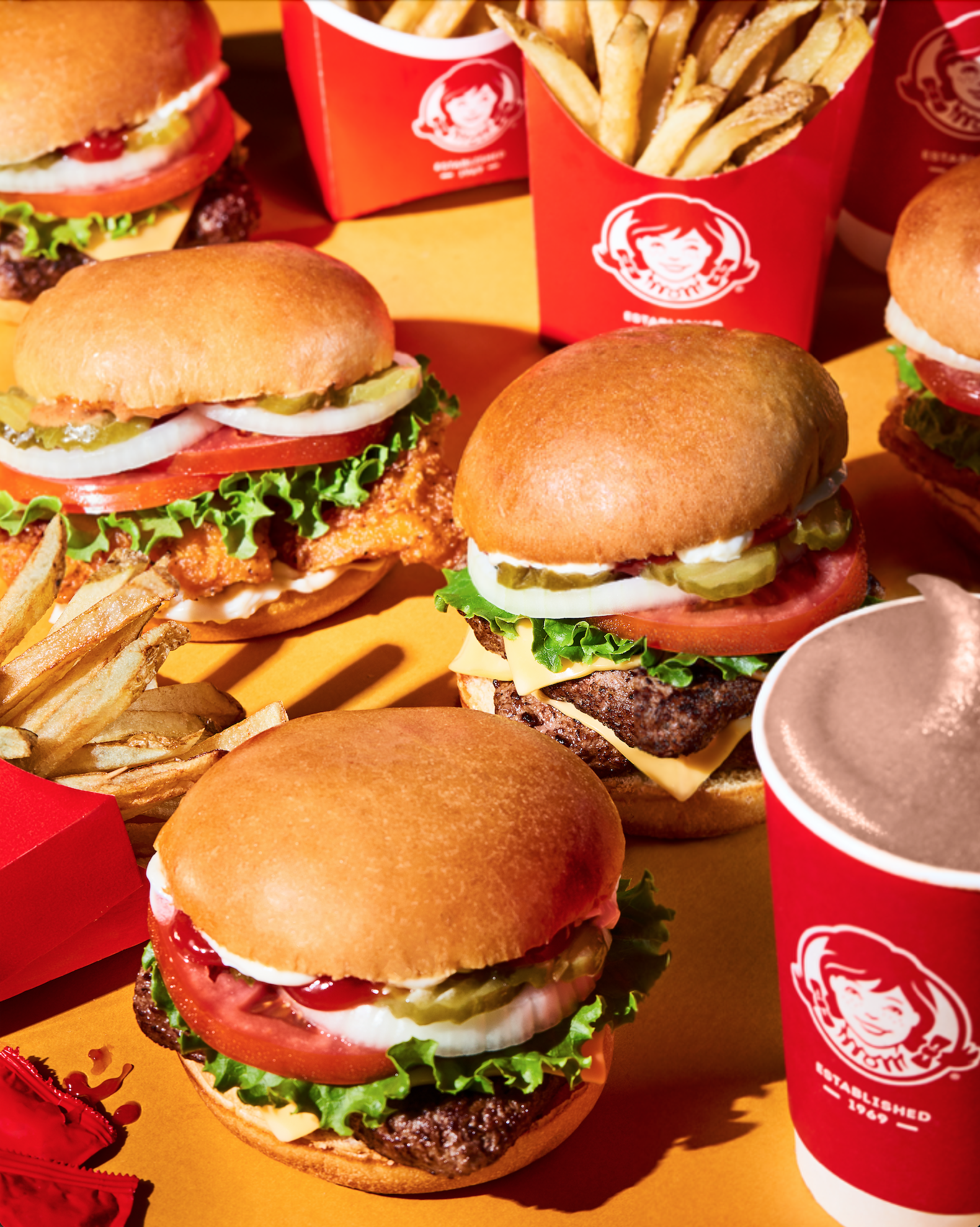 Multiple hamburgers with lettuce, tomato, pickles, cheese, and buns, along with fries and sodas on a yellow surface, Waffle House branding visible on cups.