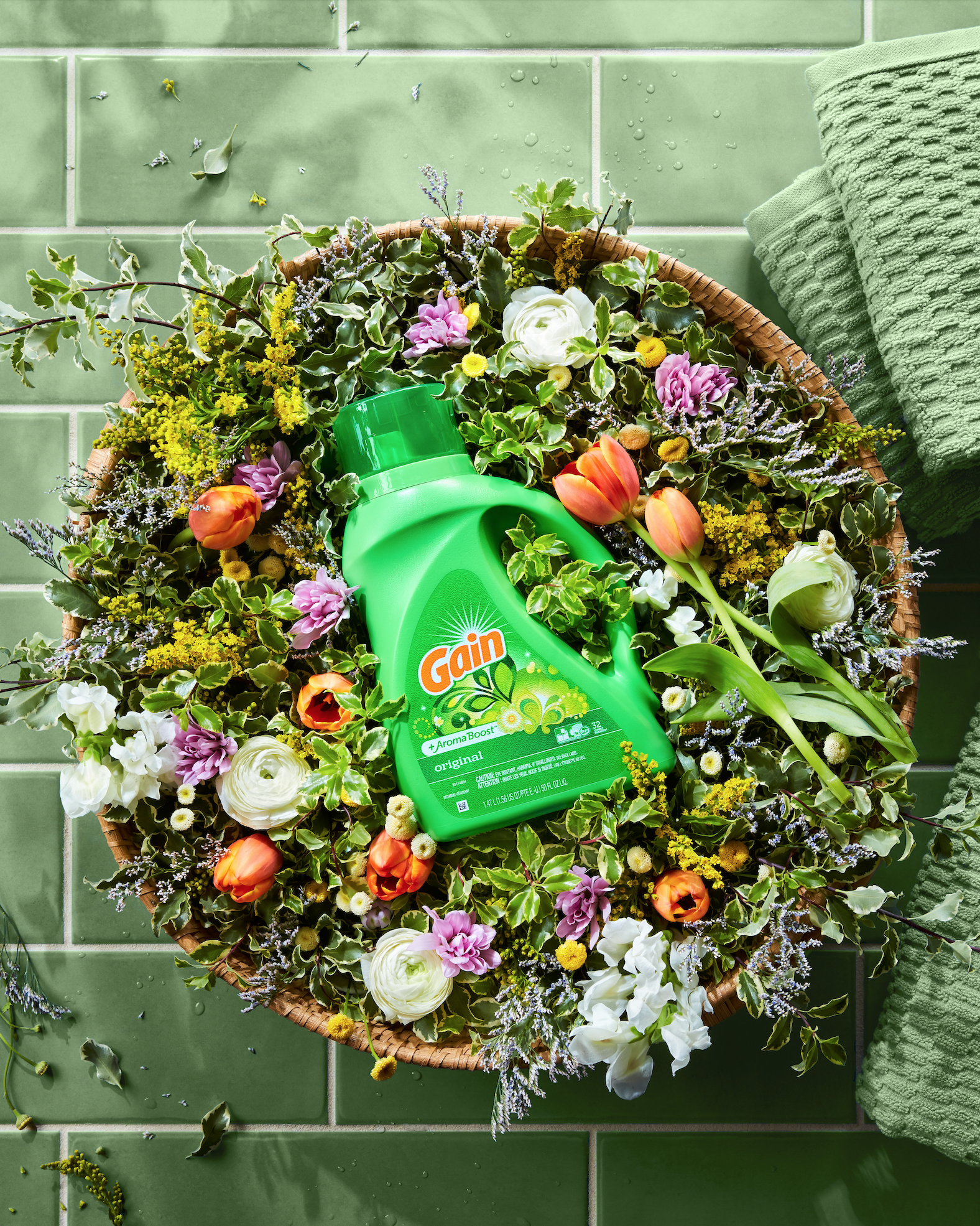 A basket of colorful flowers with a bottle of Gain laundry detergent placed in the center on a green tiled floor with green towels nearby.