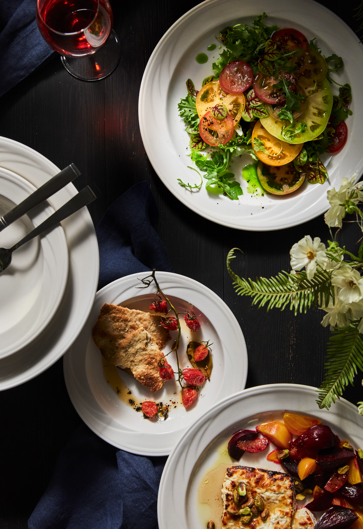 A banquet table with various dishes including a colorful tomato salad, a slice of breaded dish with cherry tomatoes, and a dish with cheese and roasted vegetables, alongside a glass of red wine and flowers.