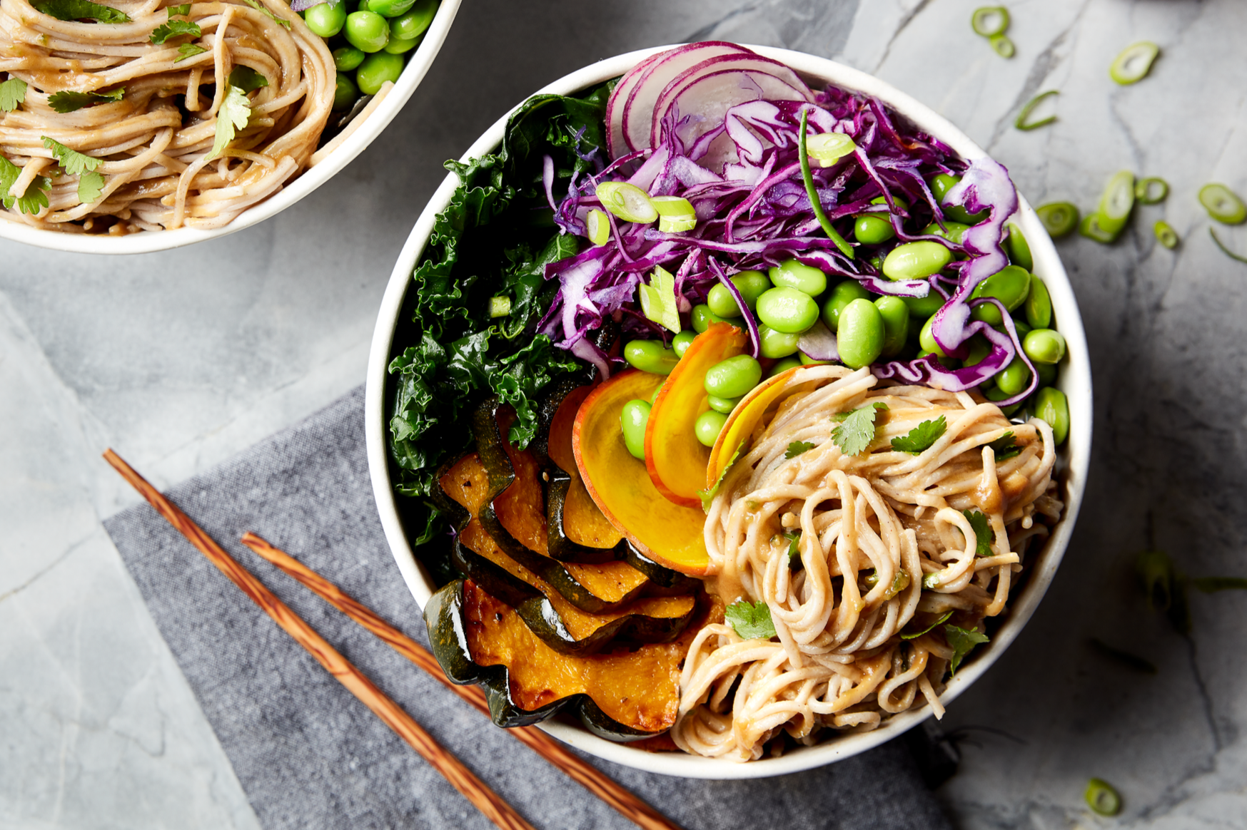 A bowl of colorful mixed salad with shredded purple cabbage, green peas, sliced radishes, roasted pumpkin and yellow squash, and cooked noodles garnished with cilantro, on a gray marble surface with a pair of chopsticks nearby.