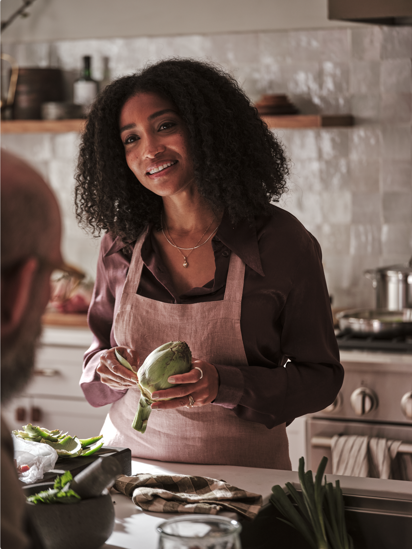 A woman wearing a beige apron and a dark shirt is smiling and holding a purple artichoke in a kitchen with gray tiles and wooden shelves.