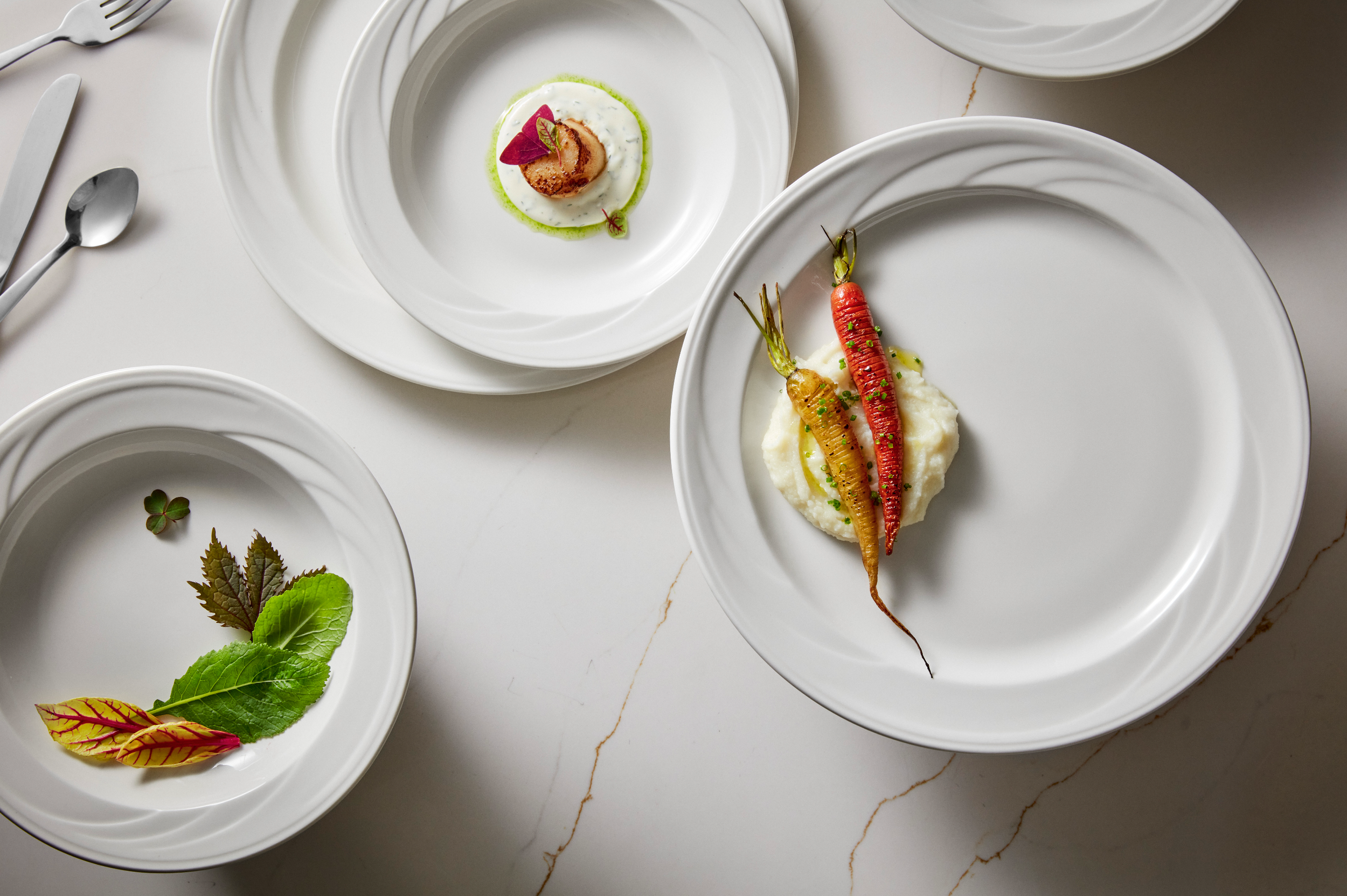 Three white plates with gourmet food: one with carrots on mashed potatoes, another with a single scallop, and a third with leaves and herbs, all arranged elegantly on a white marble surface.