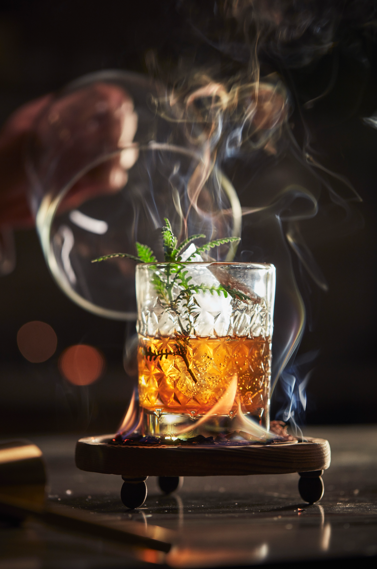 A glass containing a cocktail garnished with a sprig of fern, with smoke rising from the drink, set on a wooden stand on a dark surface.