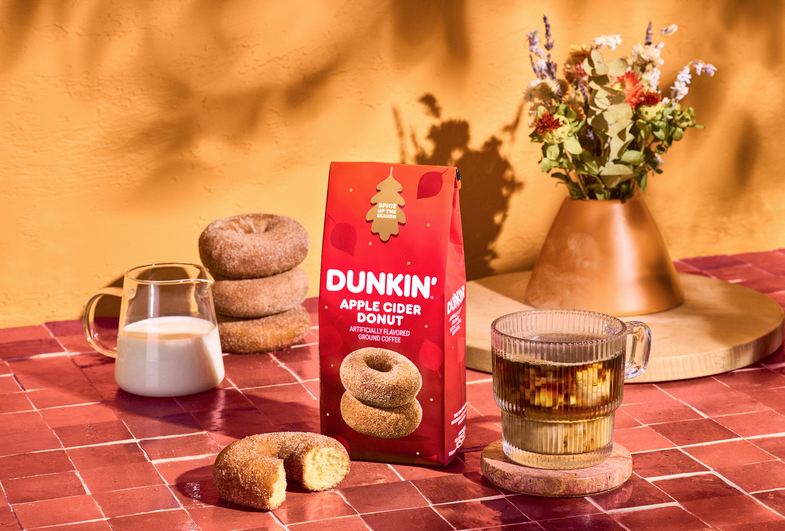 A table with a red box of Dunkin' Apple Cider Donut grounds, a glass of milk, a chocolate donut with a bite taken out of it, a glass of apple cider on a coaster, a small stack of three cinnamon sugar donuts, and a flower arrangement in a gold vase ag