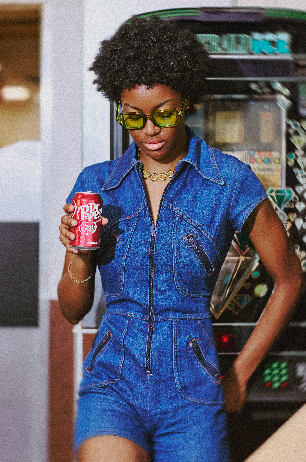 A woman wearing a denim jumpsuit and yellow sunglasses holding a can of Dr. Pepper in a vending machine area.