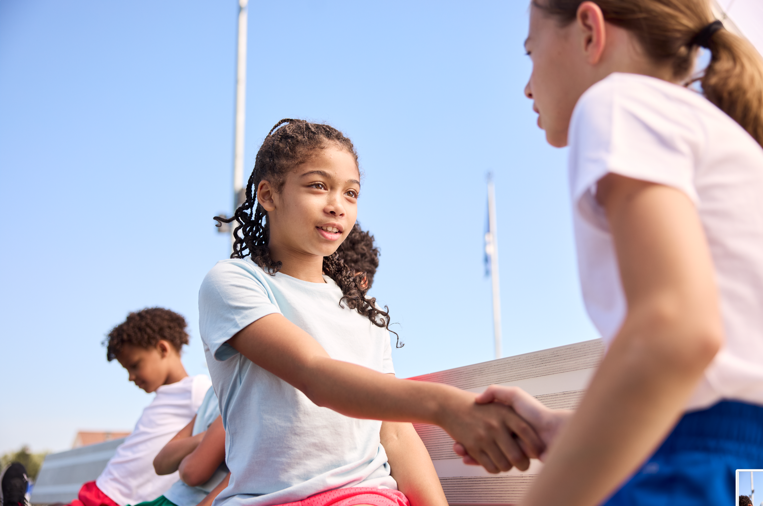 A girl shakes hands with a woman outdoors during the daytime, with other children sitting on a bench in the background.