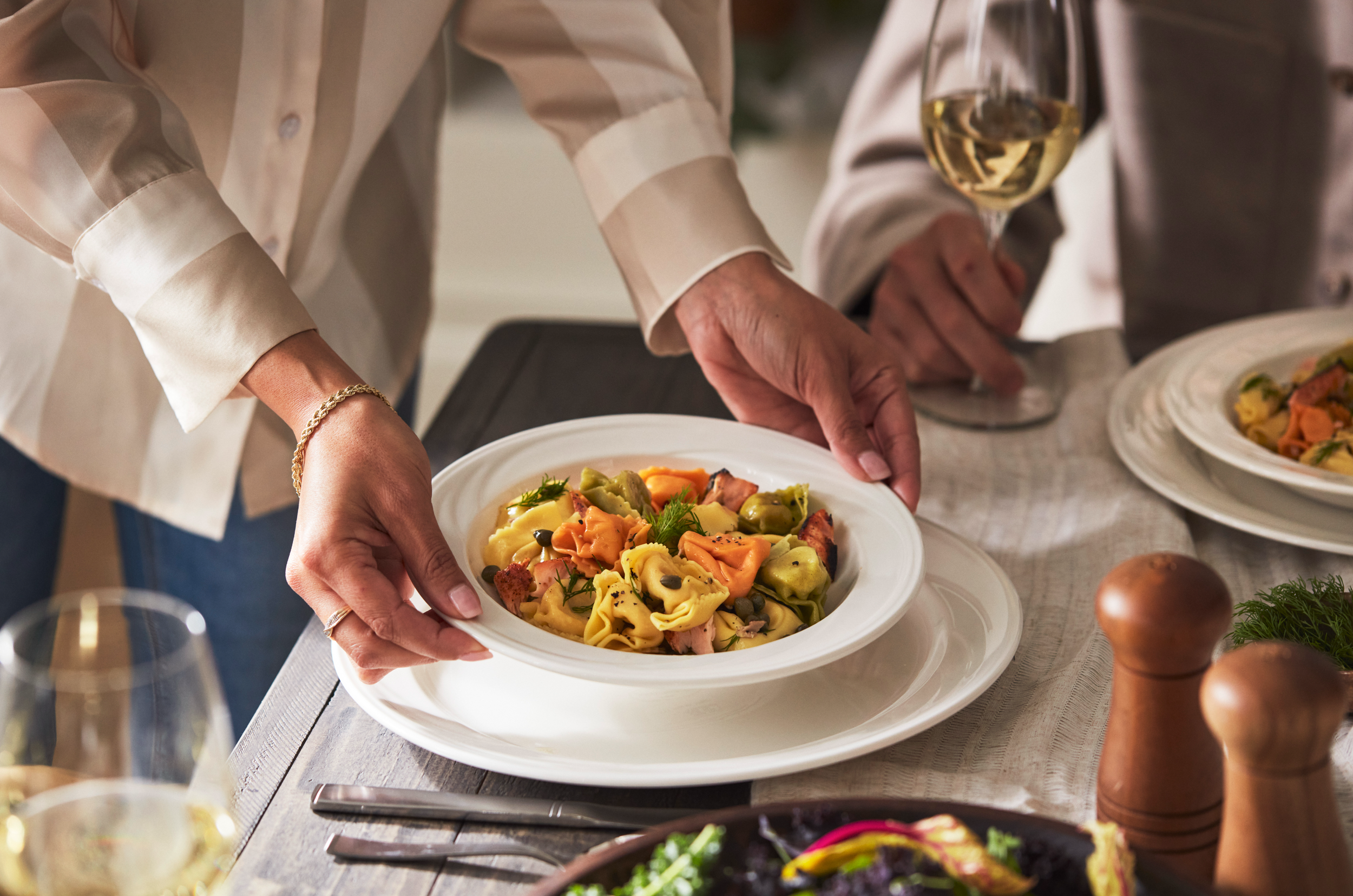 Person placing a plate of colorful tortellini pasta with herbs on a dining table with wine glasses and other dishes.