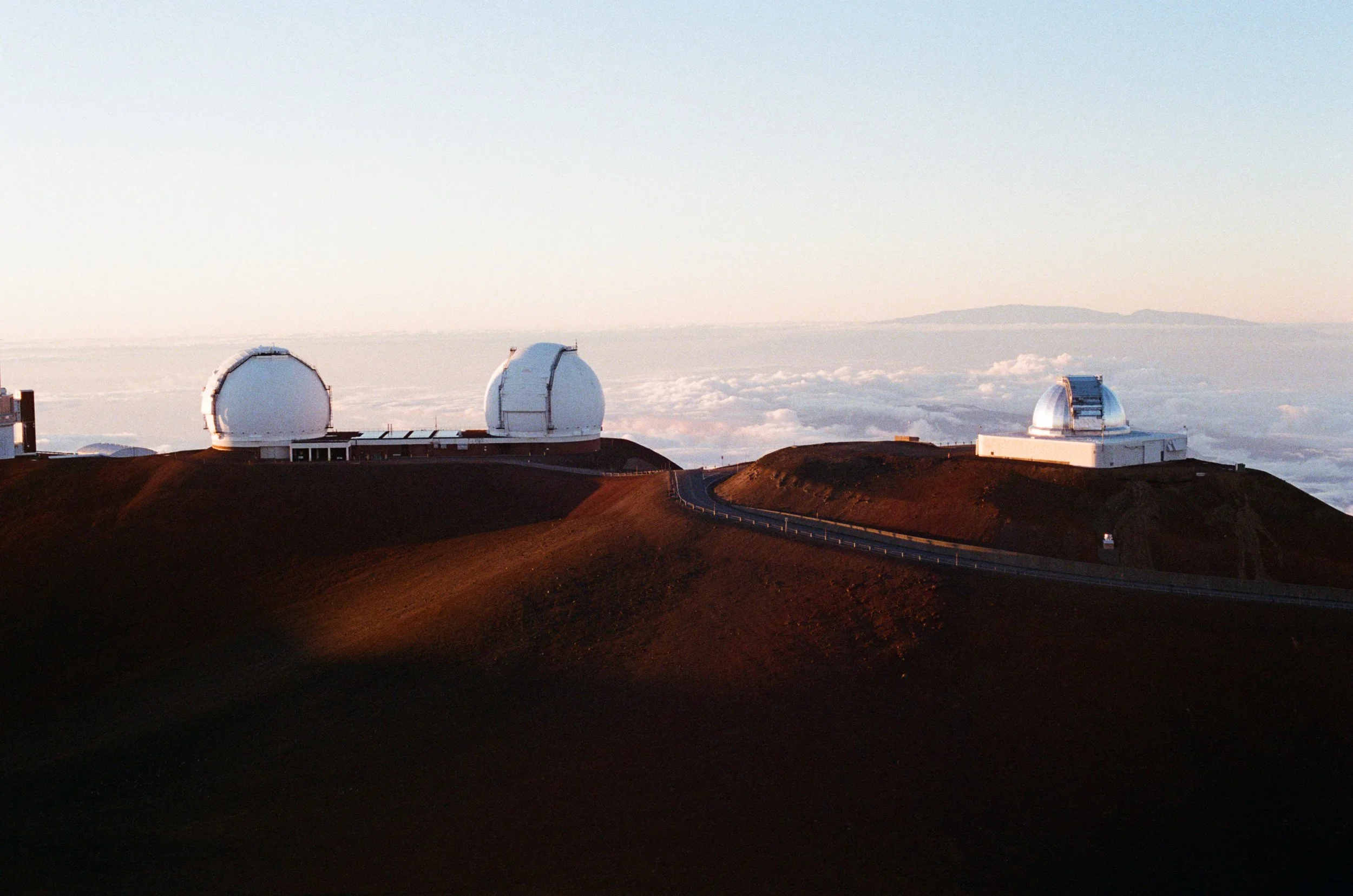 Three white observatory domes with a winding road on a mountain at sunset or sunrise, above the clouds.