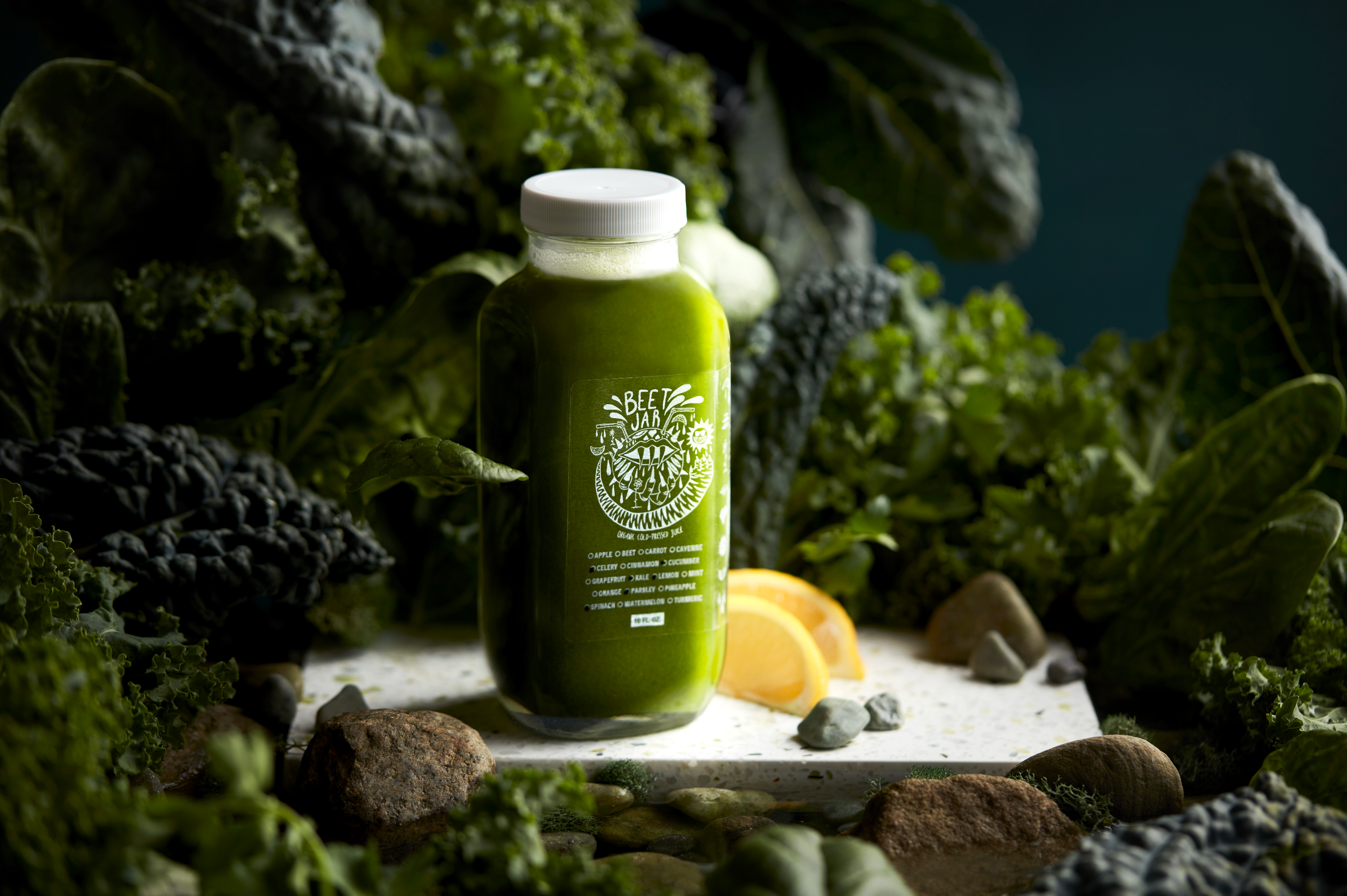 A green juice bottle labeled 'Beet Jar' surrounded by leafy greens, lemon slices, and rocks on a white surface with a dark background.