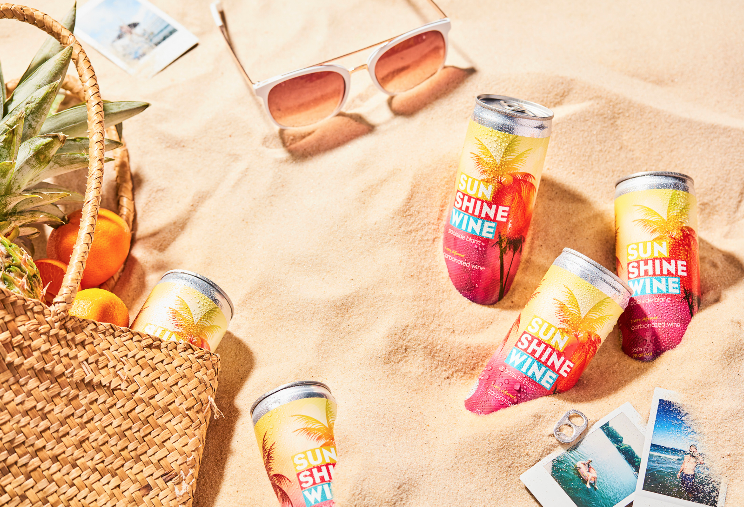 Colorful cans of Sun Shine Wine with a tropical theme scattered on sandy beach, next to a woven basket with pineapples and oranges, sunglasses, and holiday photos.