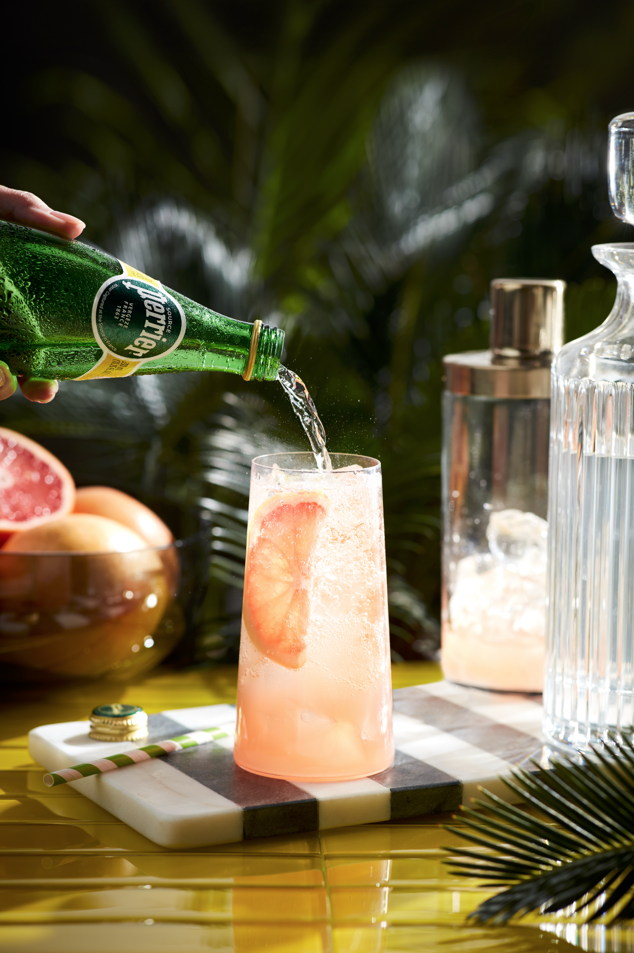 A hand pouring Perrier sparkling water into a pink grapefruit soda with ice and a grapefruit slice, on a black and white marble coaster, with a bowl of peaches and grapefruit in the background, and tropical palm leaves.
