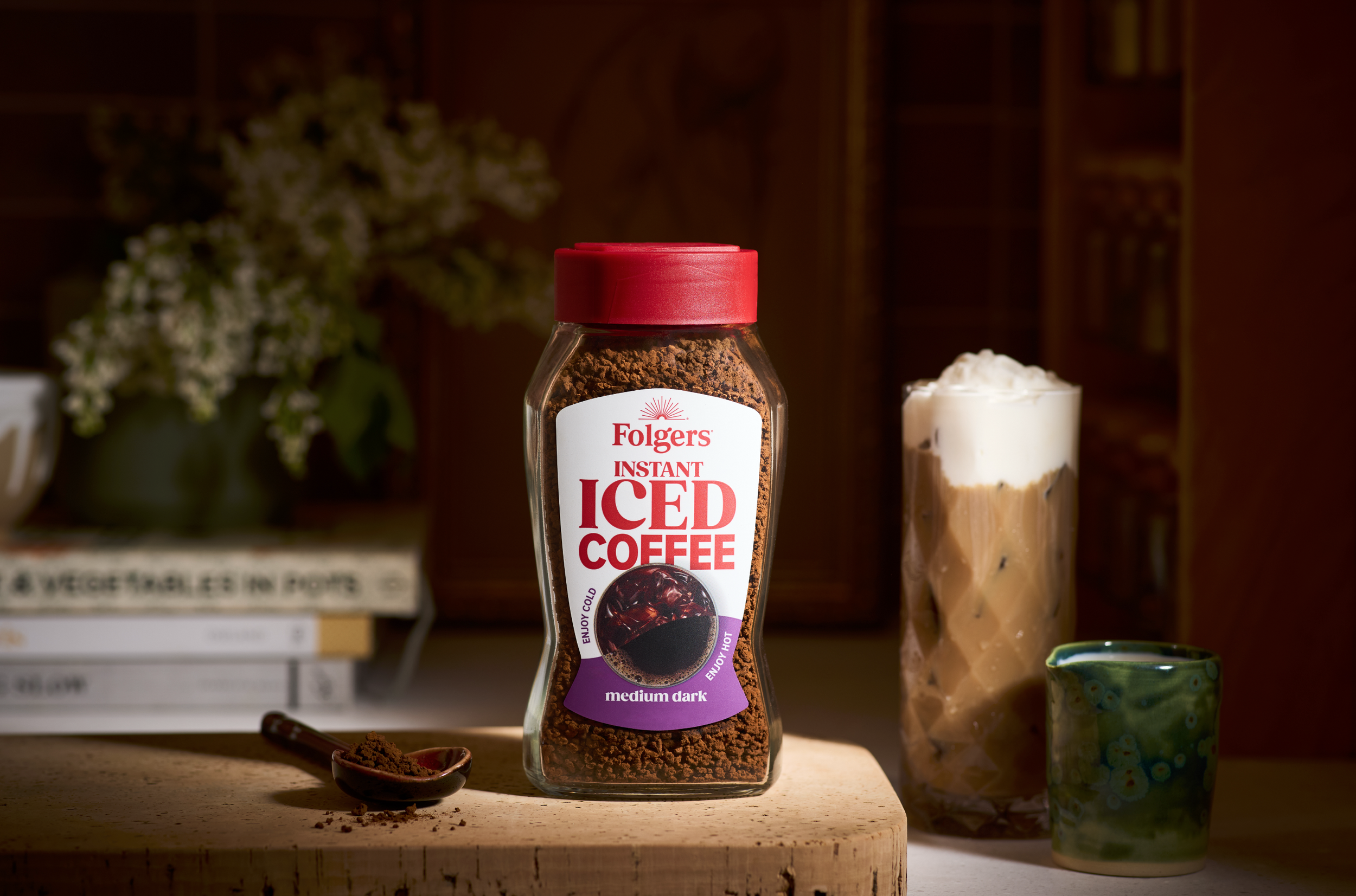 A jar of Folgers instant iced coffee powder with medium dark roast flavor on a wooden surface, a small spoon with coffee powder, and a tall glass of iced coffee topped with whipped cream in the background.