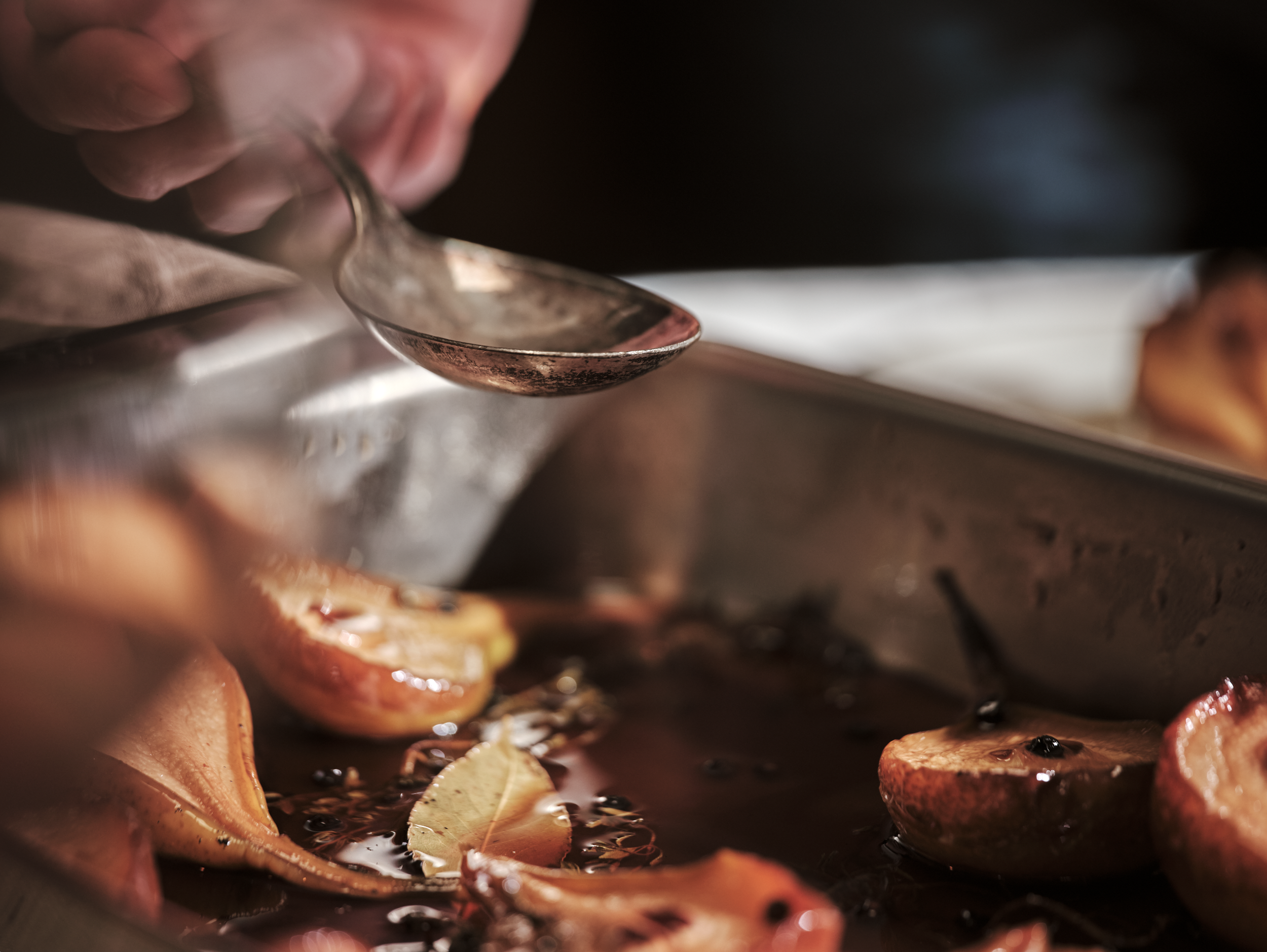 Close-up of a person using a spoon to scoop sauce or liquid from a tray of roasted or baked peaches with leaves and black peppercorns.