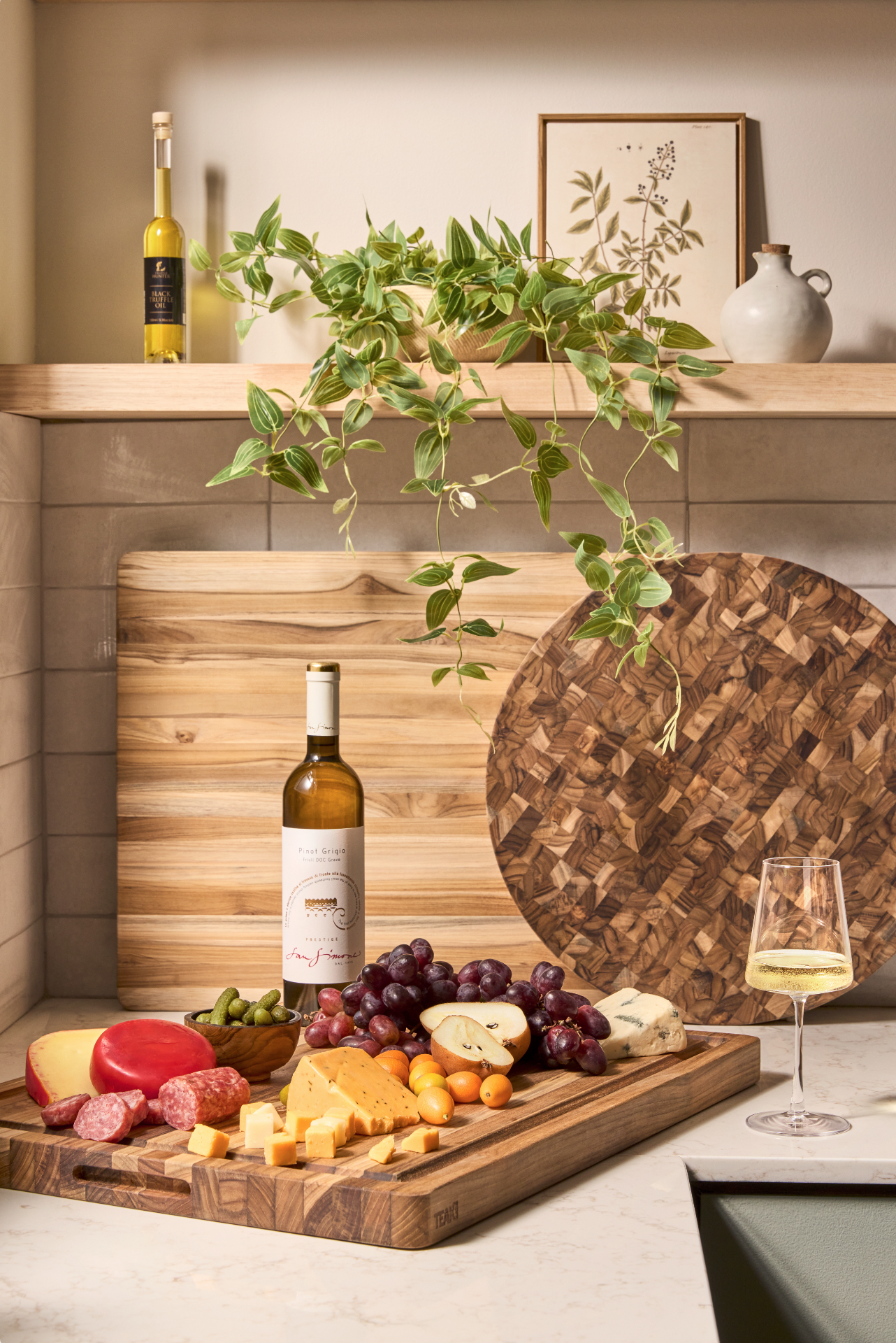 A kitchen countertop with a cheese and charcuterie board, a bottle of white wine, and a glass of wine. Green plants, wine bottles, and framed botanical artwork decorate the background.
