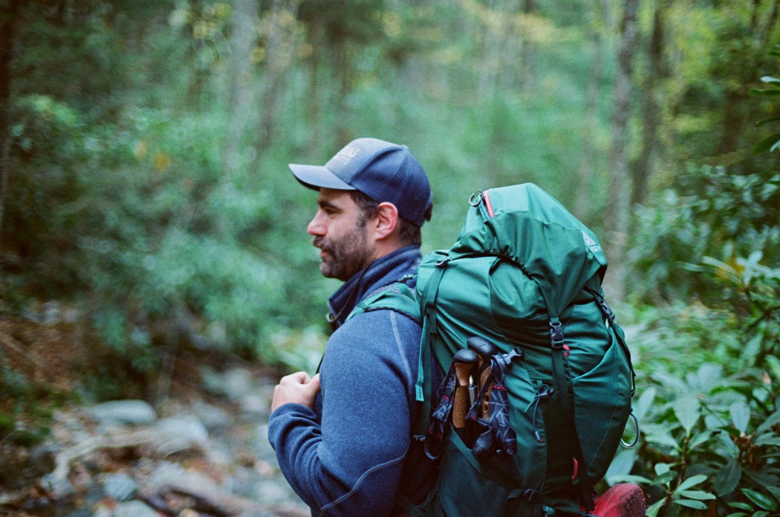 Man with a beard, wearing a blue baseball cap and a blue jacket, hiking in a green forest with a large green backpack.