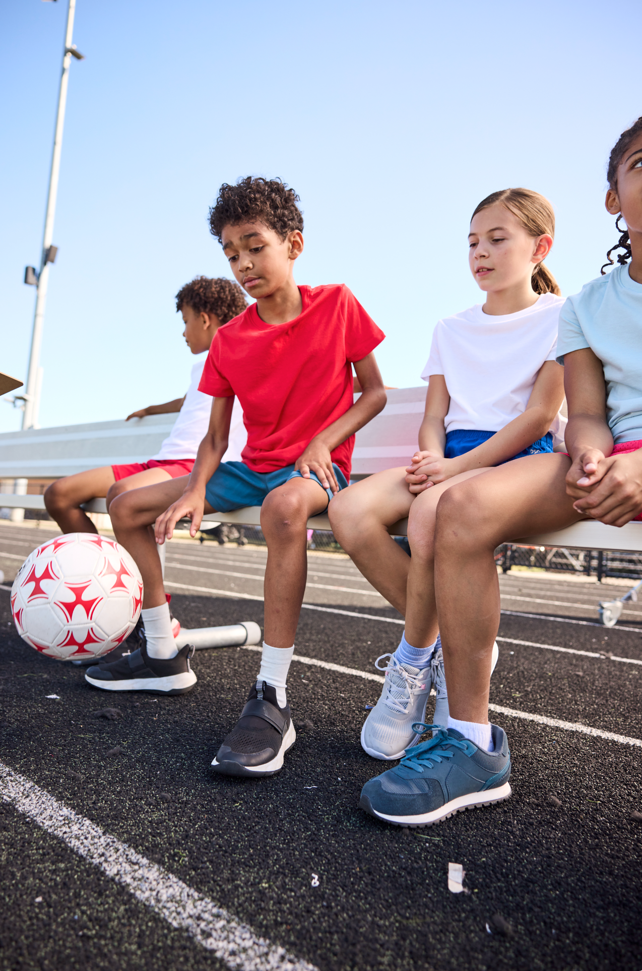 Children sitting on a bench near a running track, with one child kicking a soccer ball.