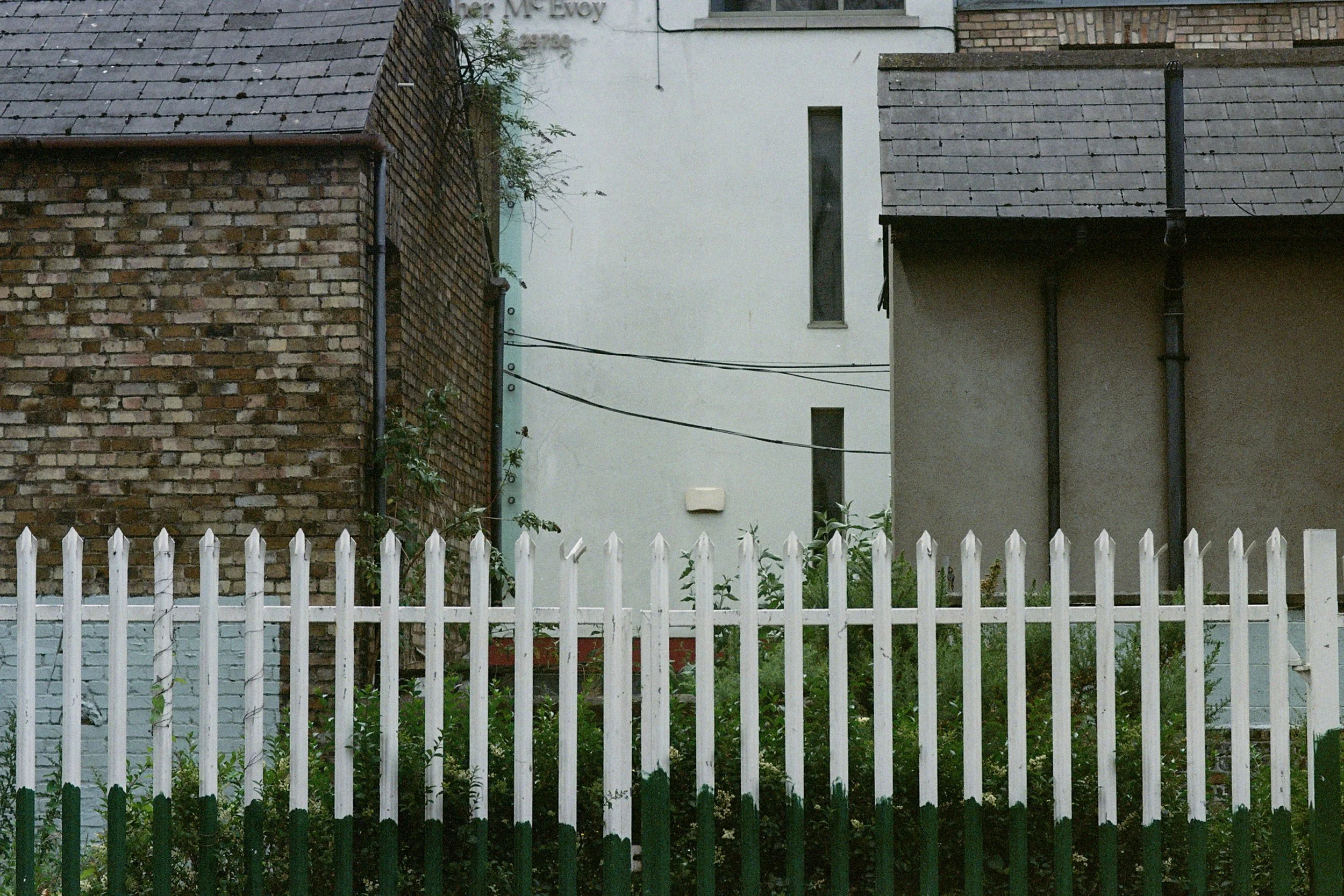 Urban scene with a white picket fence in the foreground, and three buildings with different wall textures in the background: one brick, one painted white with narrow windows, and one gray with pipes. Overcast sky.