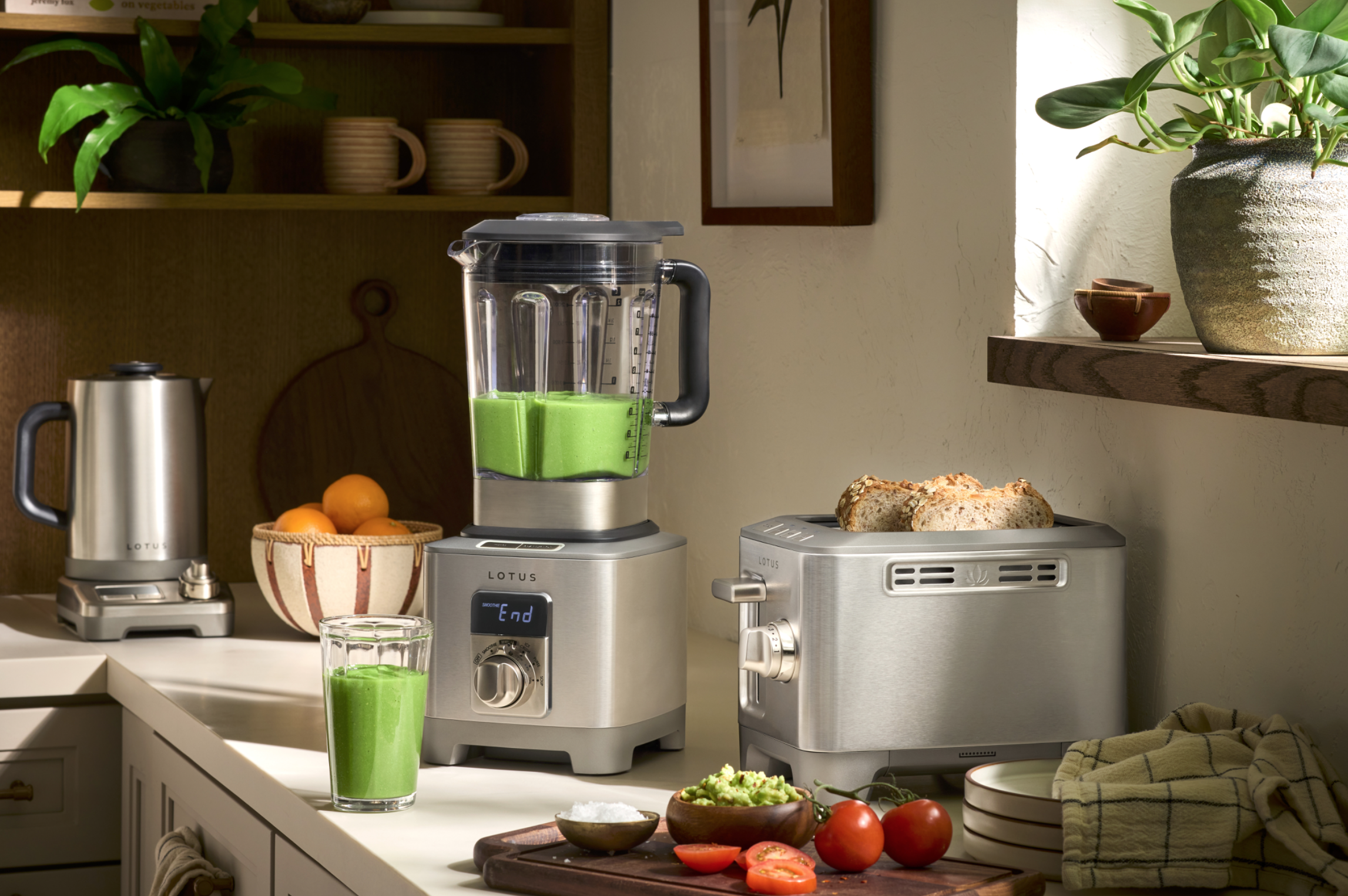 Kitchen countertop with a blender containing green smoothie, a glass of green smoothie, toast on a toaster, a bowl of oranges, tomatoes, guacamole, and sliced tomatoes, with a bowl of processed ingredients and a towel nearby.