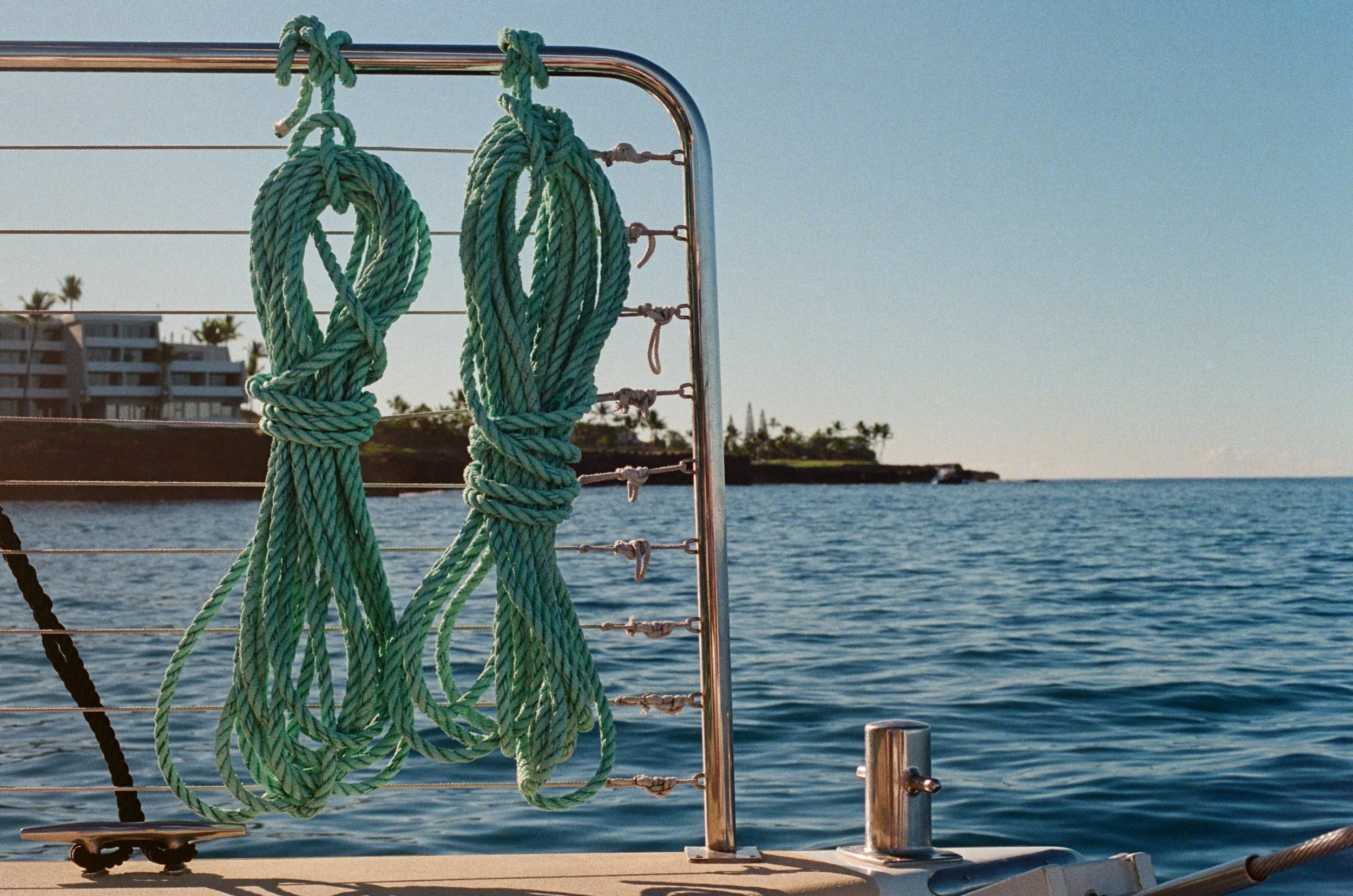 Two thick green ropes tied in loops hanging on a metal railing aboard a boat, with a calm ocean, distant shoreline, and clear sky in the background.