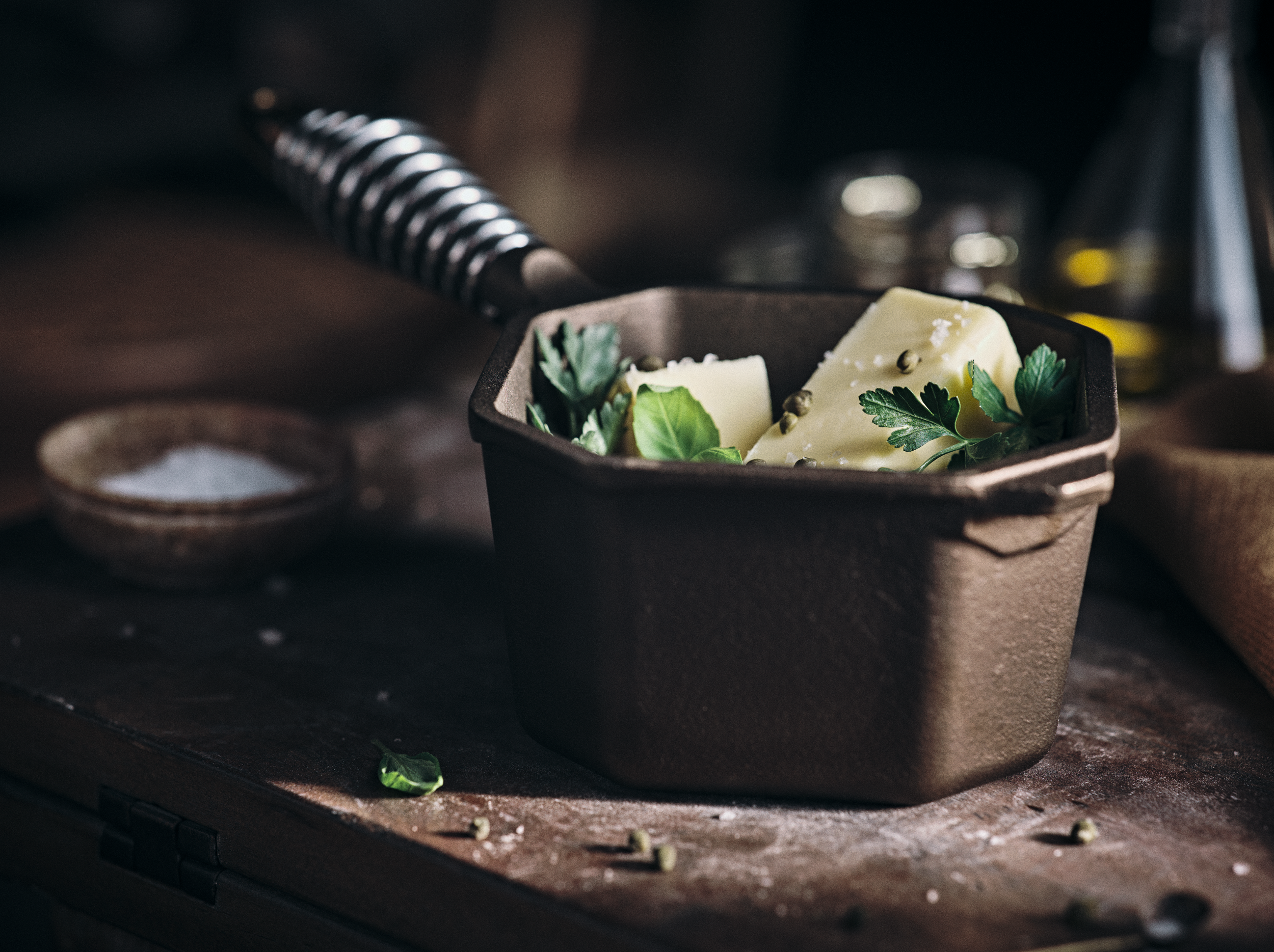 A black baking pan filled with two slices of cheese, garnished with fresh green herbs and peppercorns, on a rustic wooden surface with a small bowl of salt and scattered peppercorns nearby.