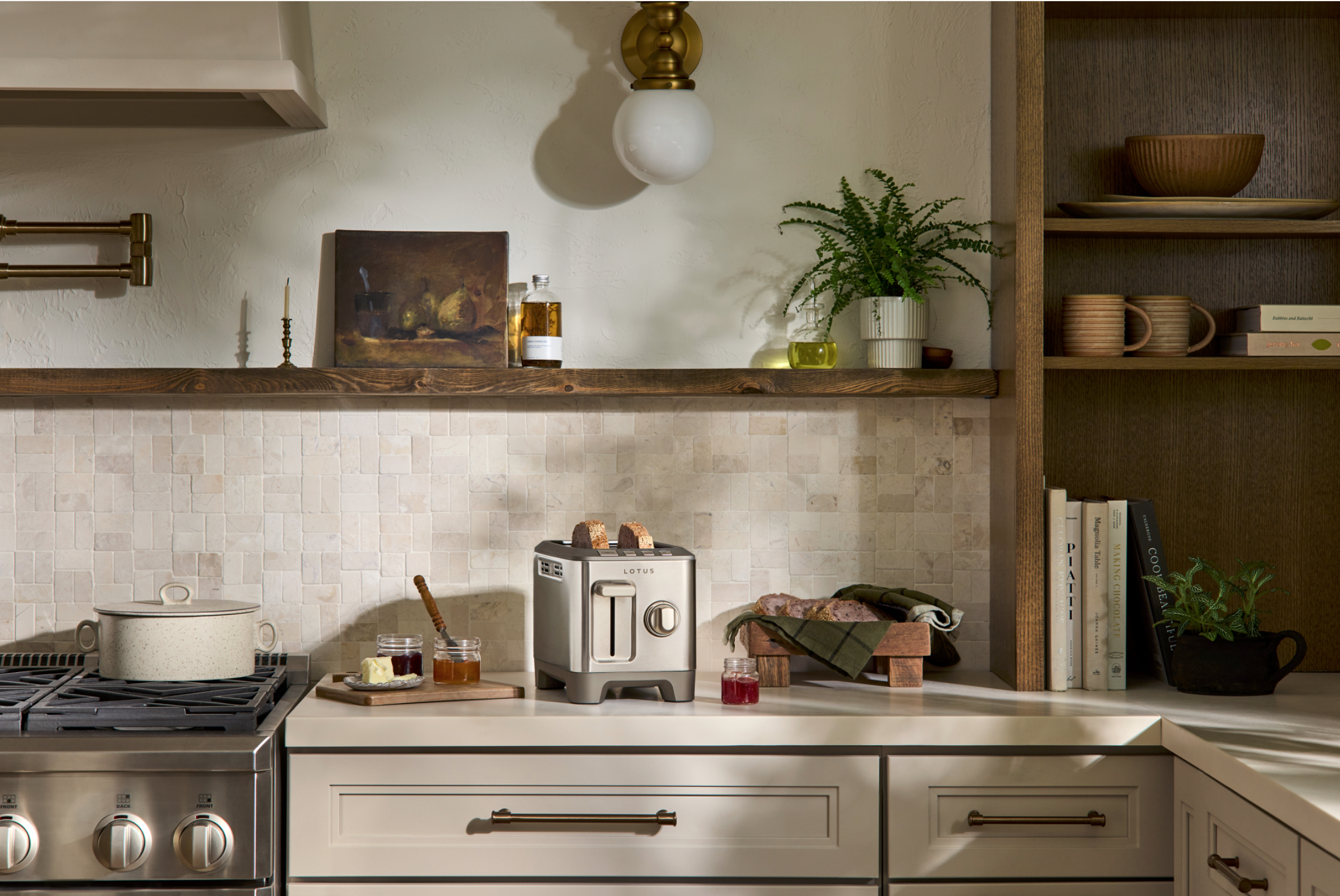 A cozy kitchen with a beige tiled backsplash, wooden shelves, and white cabinets. On the counter, there is a toaster, jars of jam, a pot with a handle, some food, and a cloth. The open shelf has books, bowls, mugs, and a potted plant. A hanging light