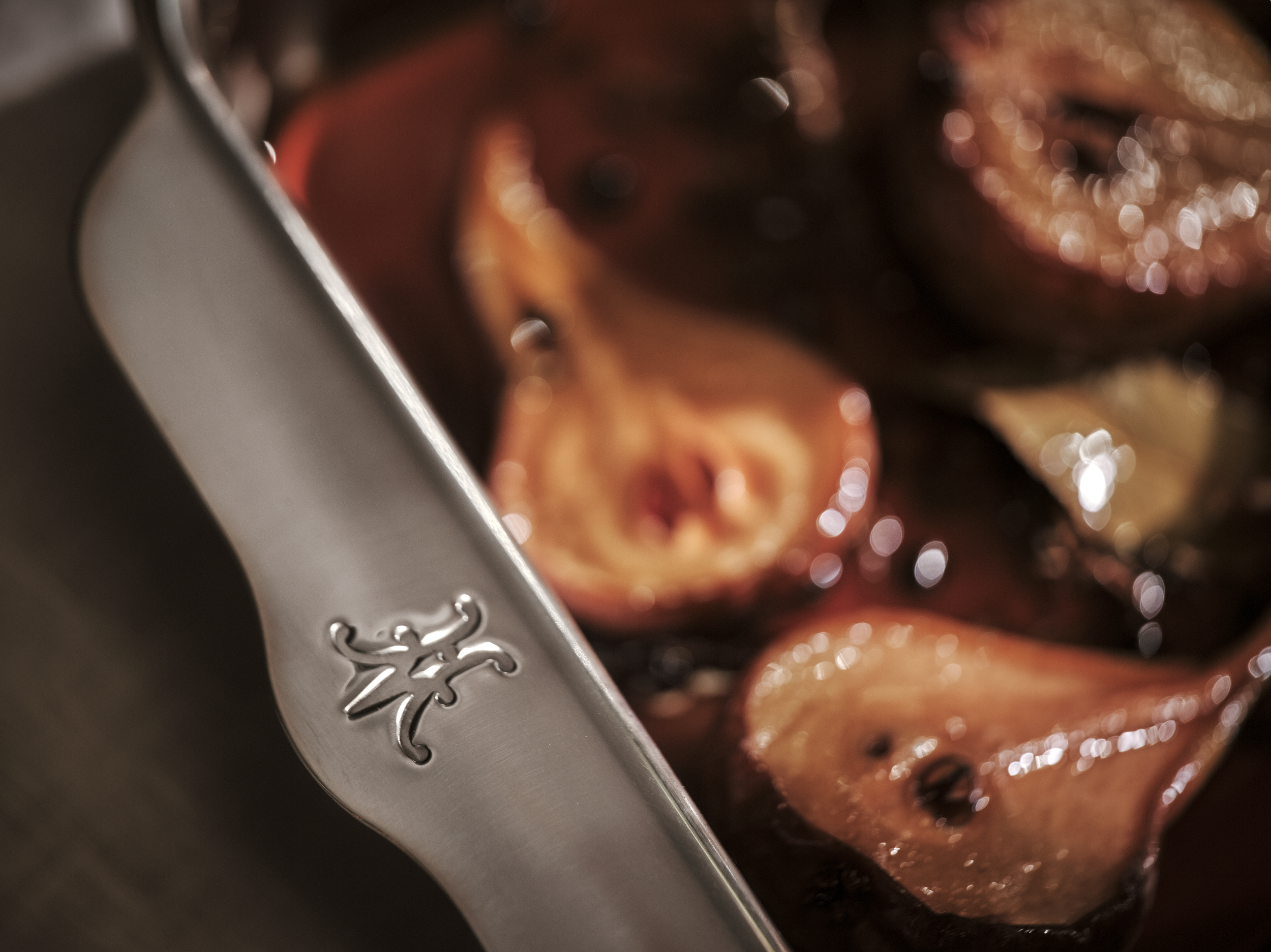 Close-up of a stainless steel chef's knife with an engraved logo, positioned next to roasted tomatoes and garlic cloves in a pan.