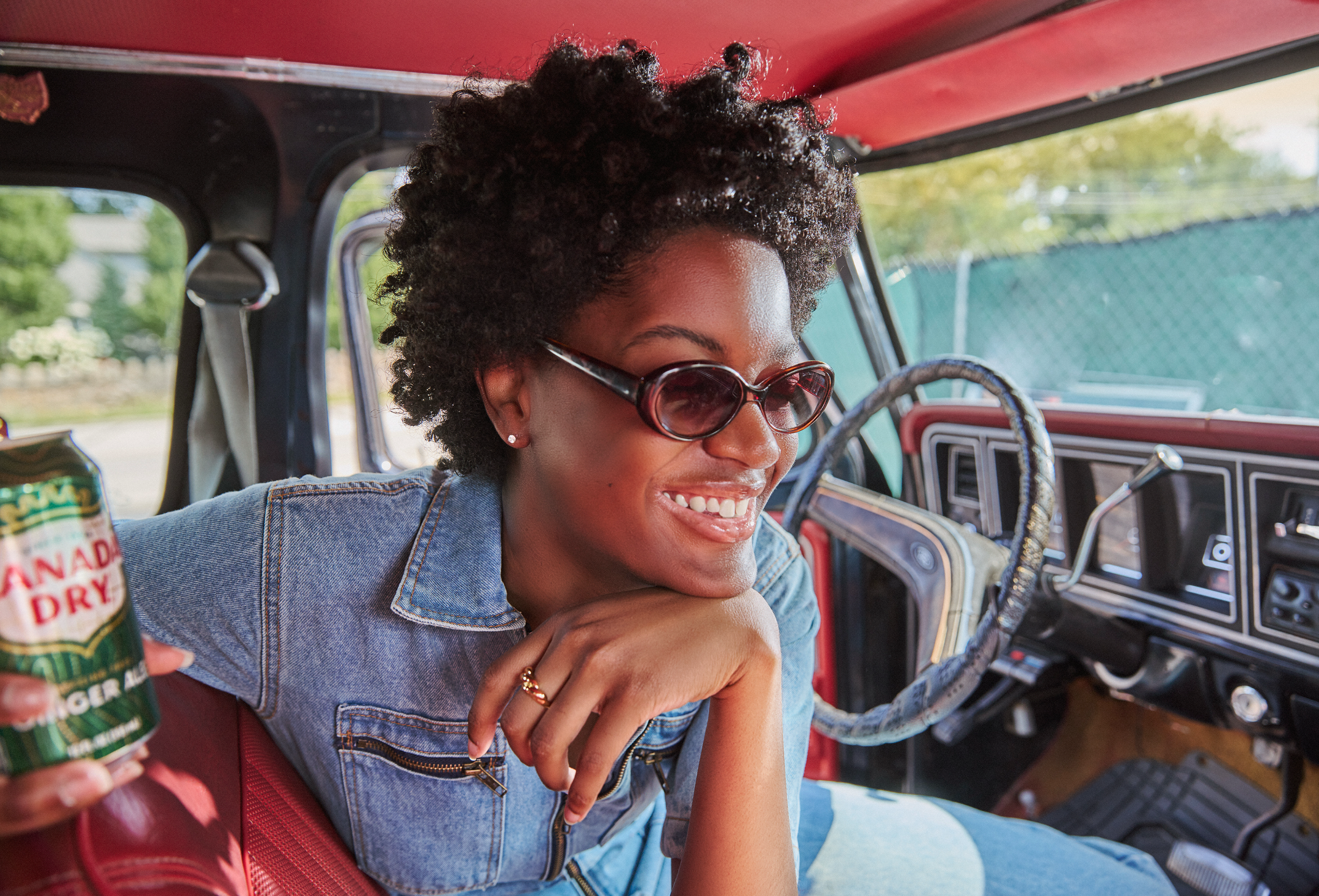 A young woman with curly hair, wearing sunglasses and a denim jacket, smiling inside an old vehicle, holding a can of Canada Dry ginger ale.