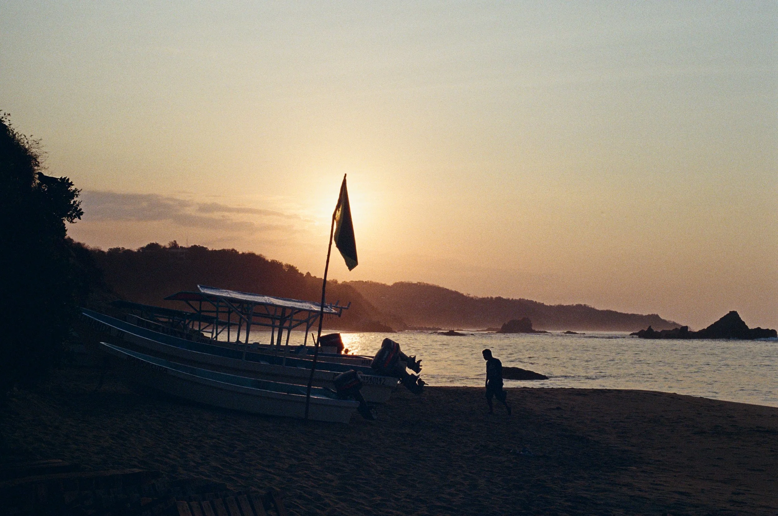 Silhouetted boats and a person walking on a beach at sunset