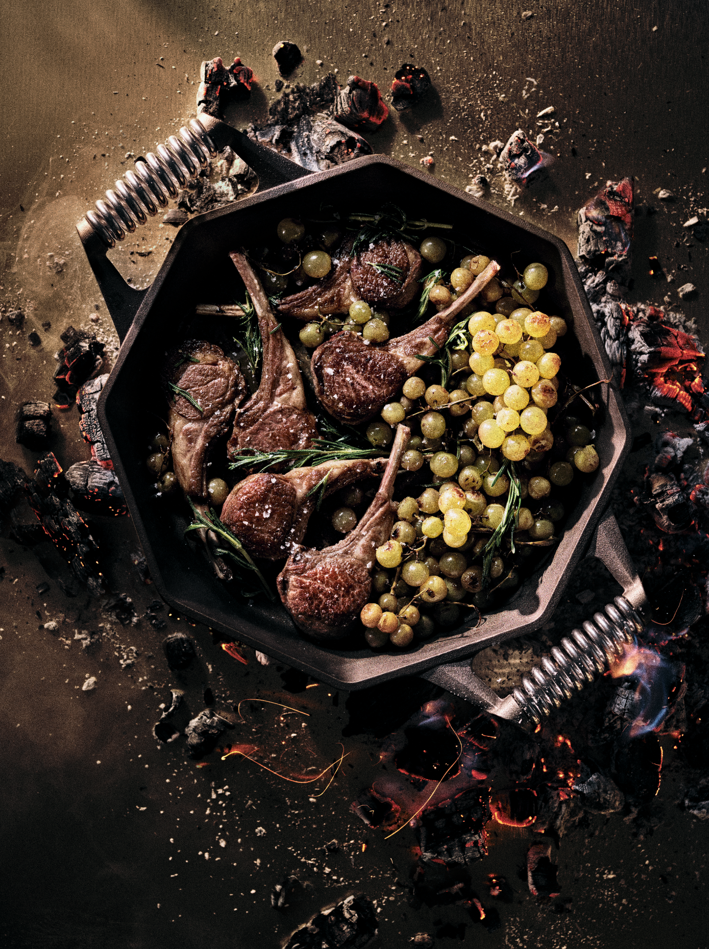 Grilled lamb chops with grapes and herbs in a black pan, surrounded by burning charcoal embers.