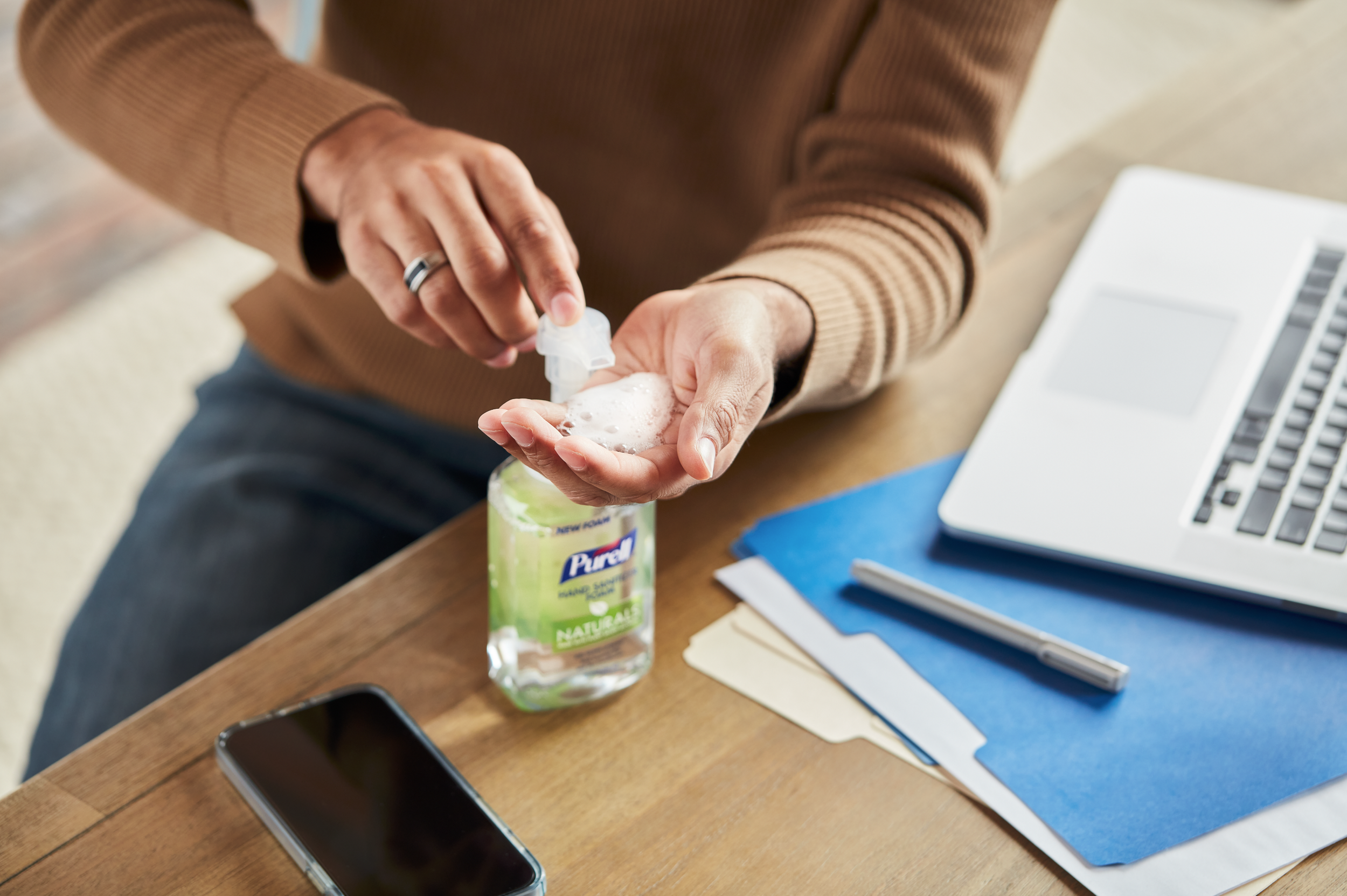 Person using hand sanitizer on their palm at a workspace with a laptop, smartphone, folders, and papers.