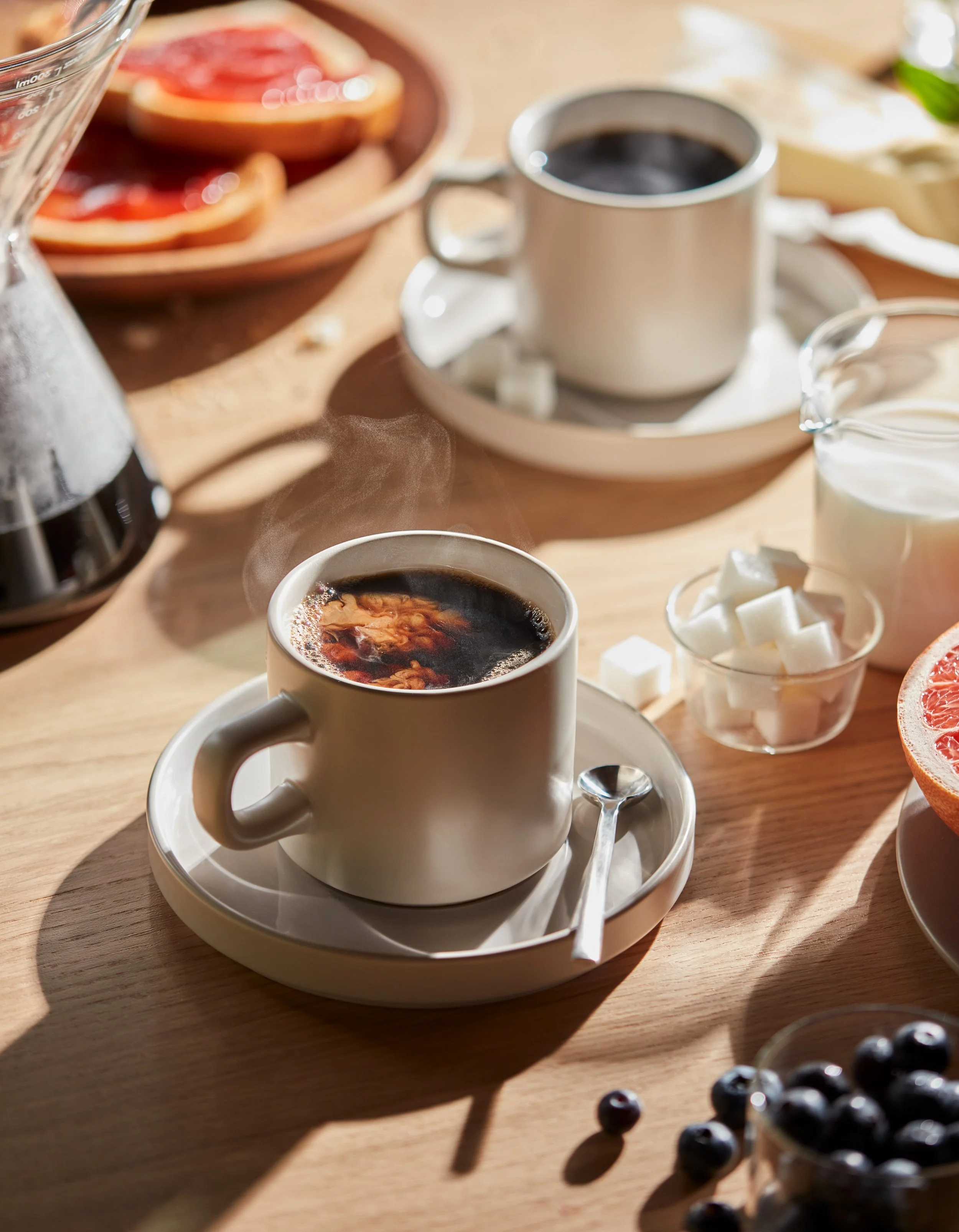 A cup of steaming black coffee on a wooden table surrounded by breakfast items, including sugar cubes, grapefruit slices, and a pitcher of cream.