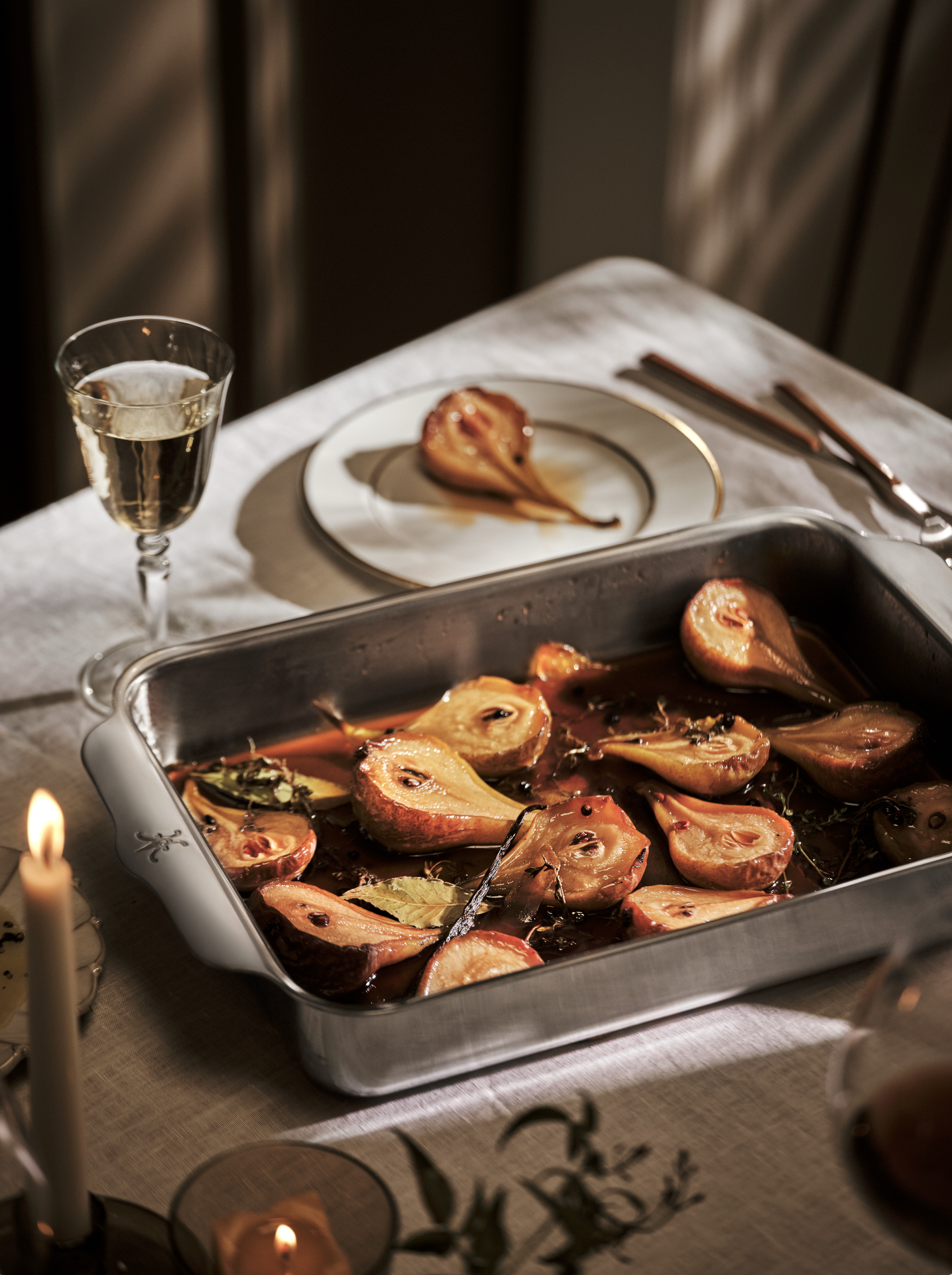 A table set for a meal with roasted pears in a metallic baking dish, a glass of champagne, a plate with a roasted pear, and cutlery. Candles and a tablecloth are also visible.