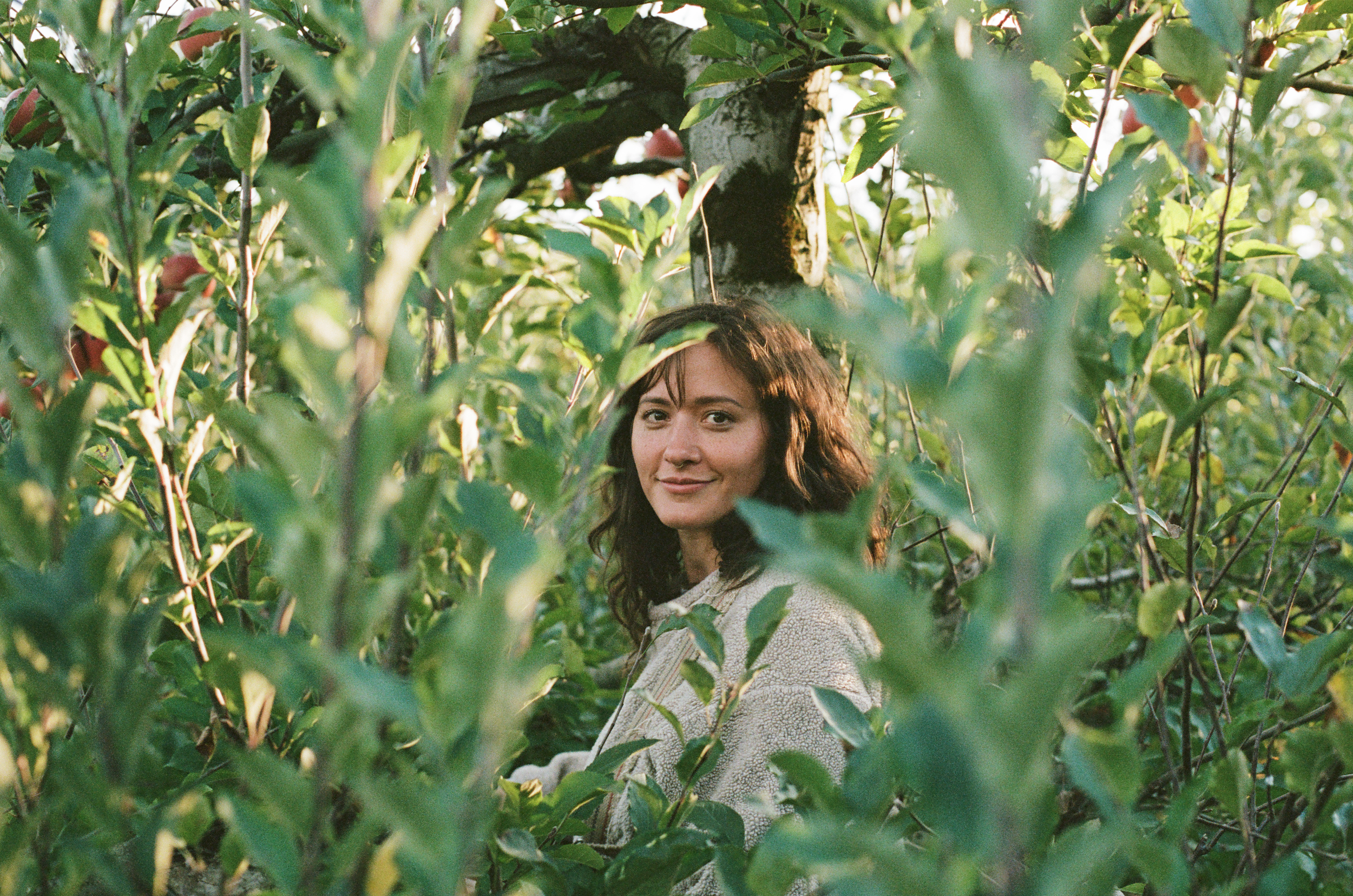 A woman with brown curly hair smiling while standing among green foliage with some red fruit visible in the background.