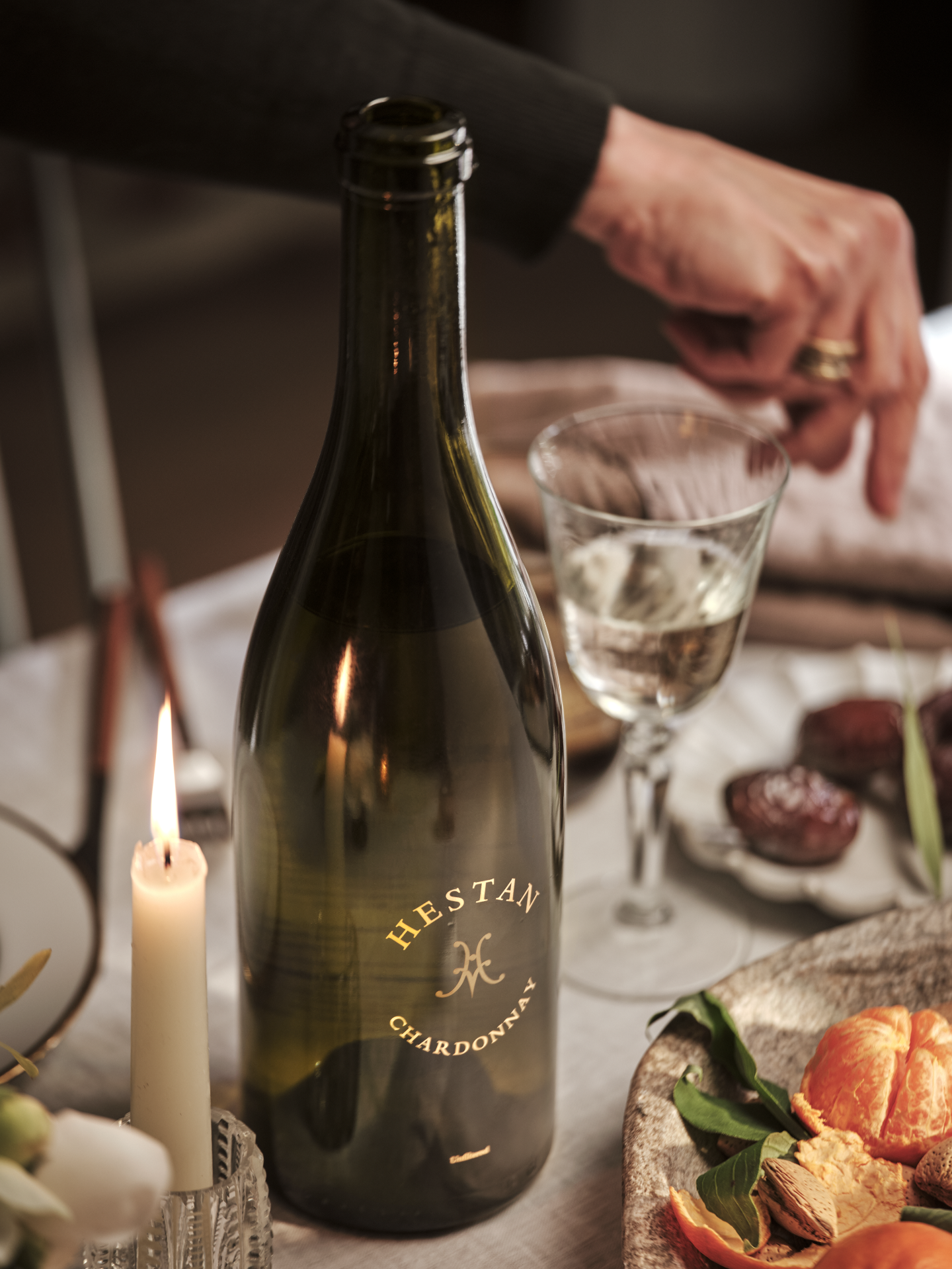 A bottle of Hestani Chardonnay wine on a dining table with a lit candle, a glass of wine, and a plate of dried fruits and nuts, with a person's hand reaching for something in the background.