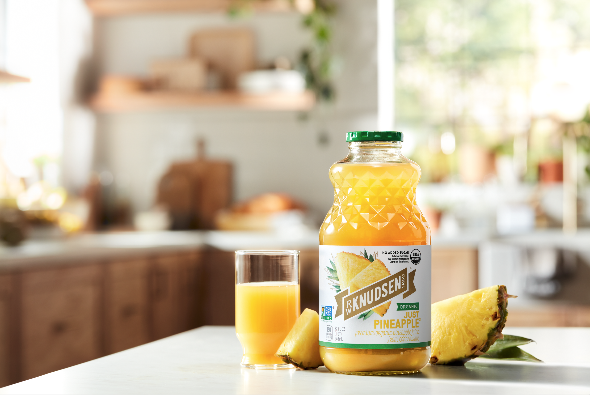 A bottle of Knudsen organic pineapple juice, a glass of pineapple juice, and a pineapple wedge on a white kitchen countertop with a blurred kitchen background.