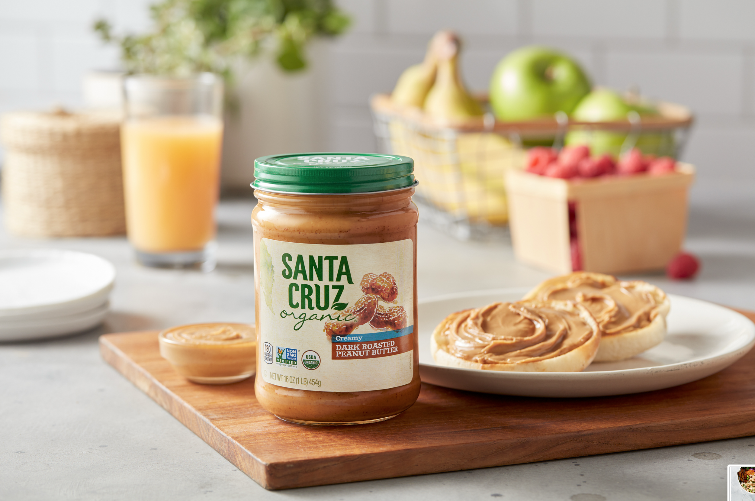 A jar of Santa Cruz organic creamy dark roasted peanut butter next to spread on bread, with a halved lemon and a plate, on a wooden board. In the background, a glass of orange juice, a basket of green apples and bananas, and a wooden box of raspberri