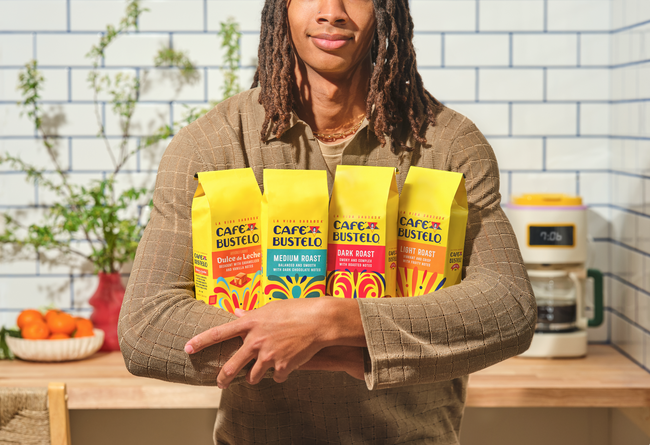 Person holding four bags of Café Bustelo coffee in a kitchen with white subway tiles, a bowl of oranges, and a coffee maker in the background.