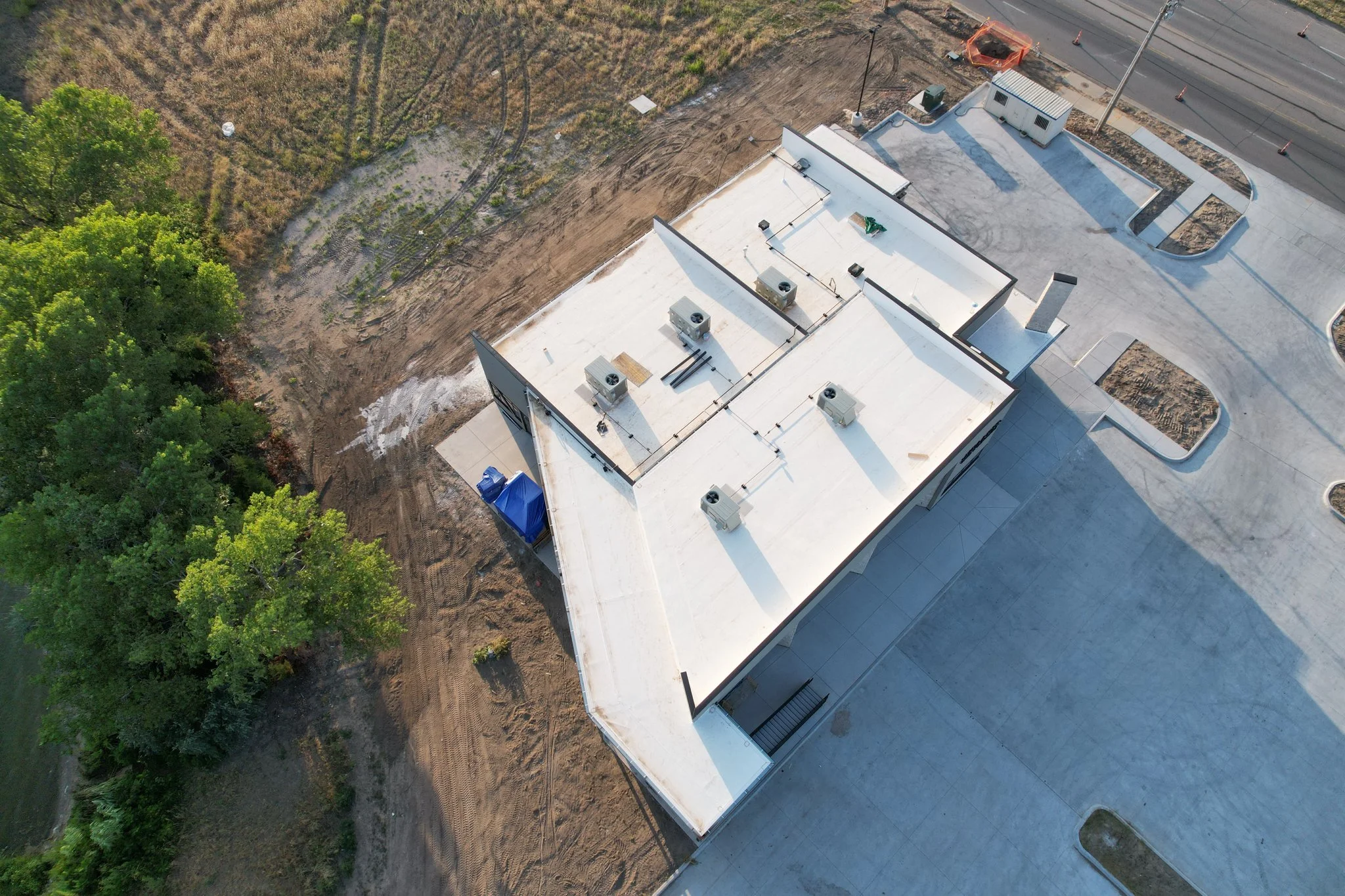 Aerial view of a commercial building with a white flat roof, surrounded by parking lot and construction area, with trees nearby.