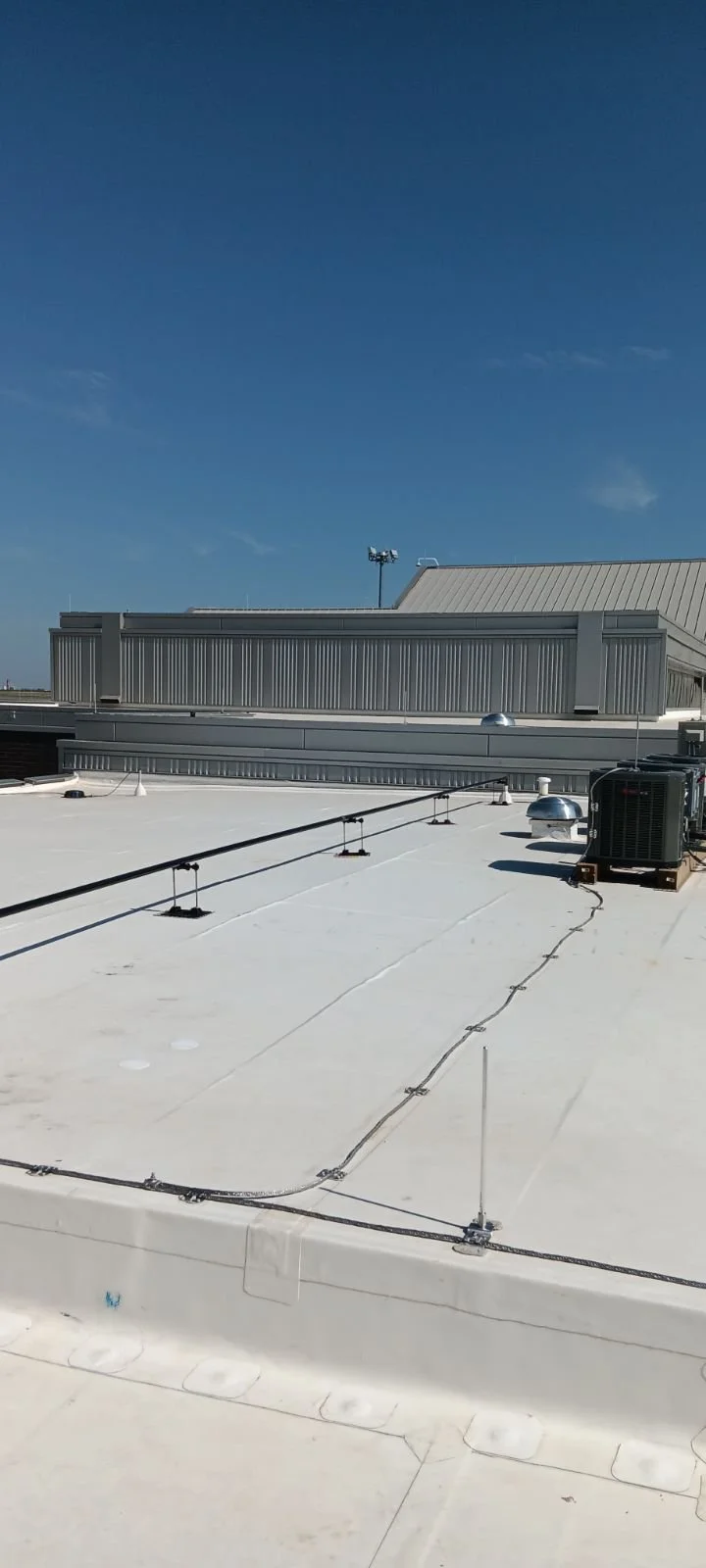 View of a flat rooftop with white surface, HVAC unit, vents, and a railing, with a blue sky and some clouds in the background.