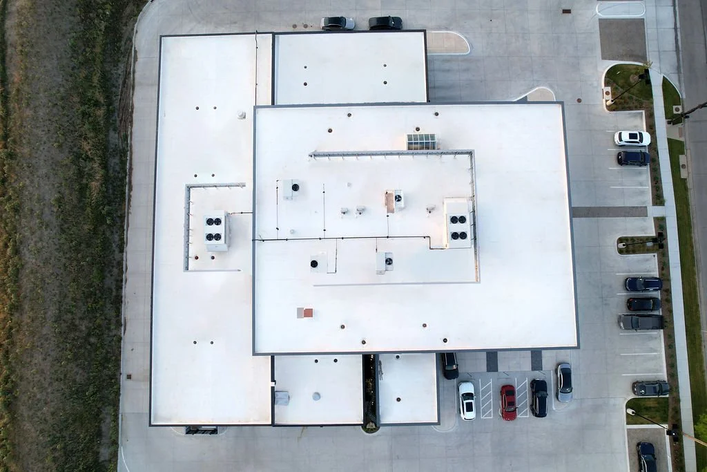 Aerial view of a building with a white flat roof, surrounded by parking lot with cars, and a small landscaped area with grass and trees.